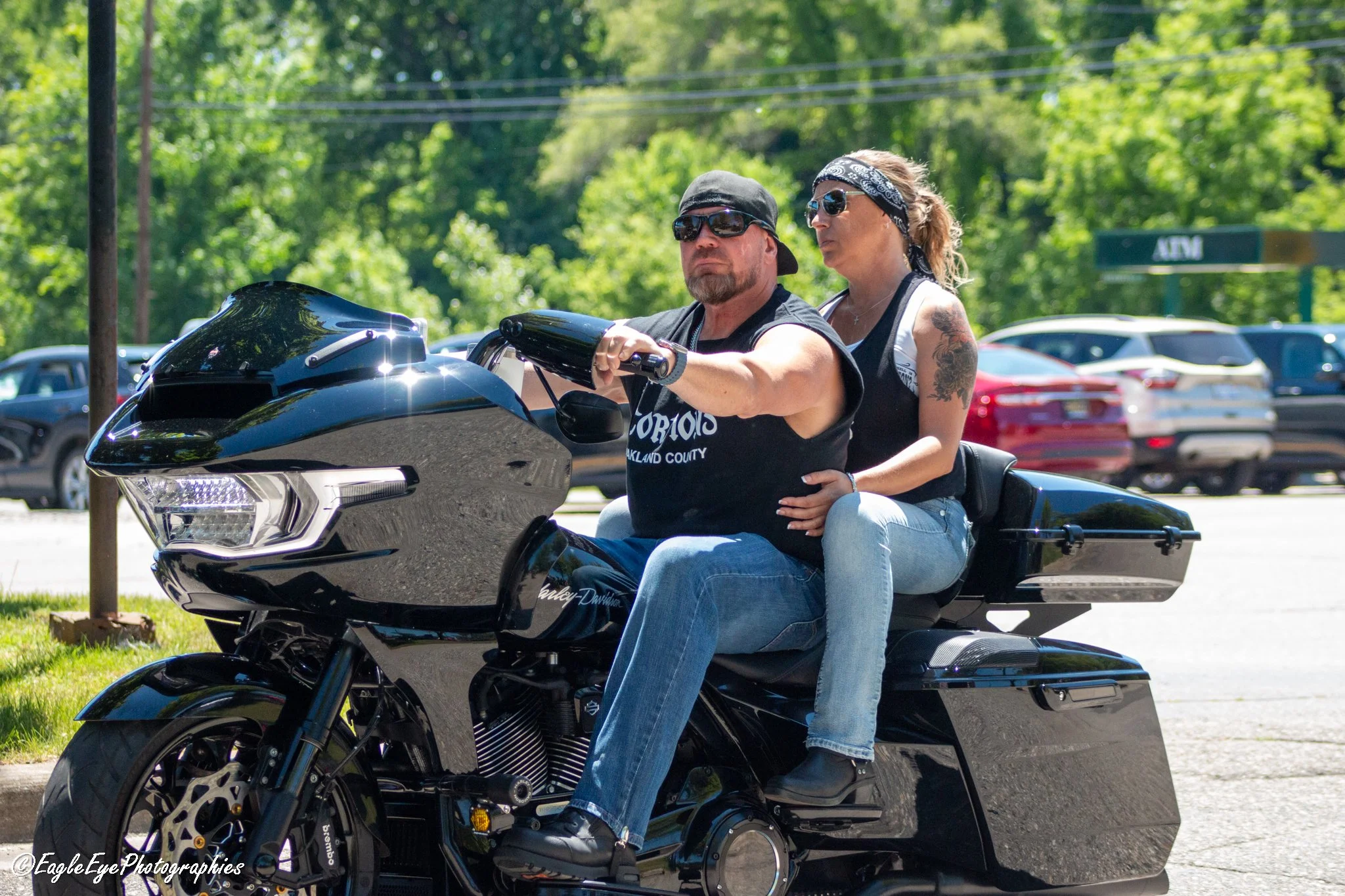 A man and woman riding a black motorcycle in a parking lot during daytime. The man is wearing sunglasses, a black bandana, and a sleeveless shirt, while the woman is seated behind him, also wearing sunglasses and a bandana, with a tattoo on her upper