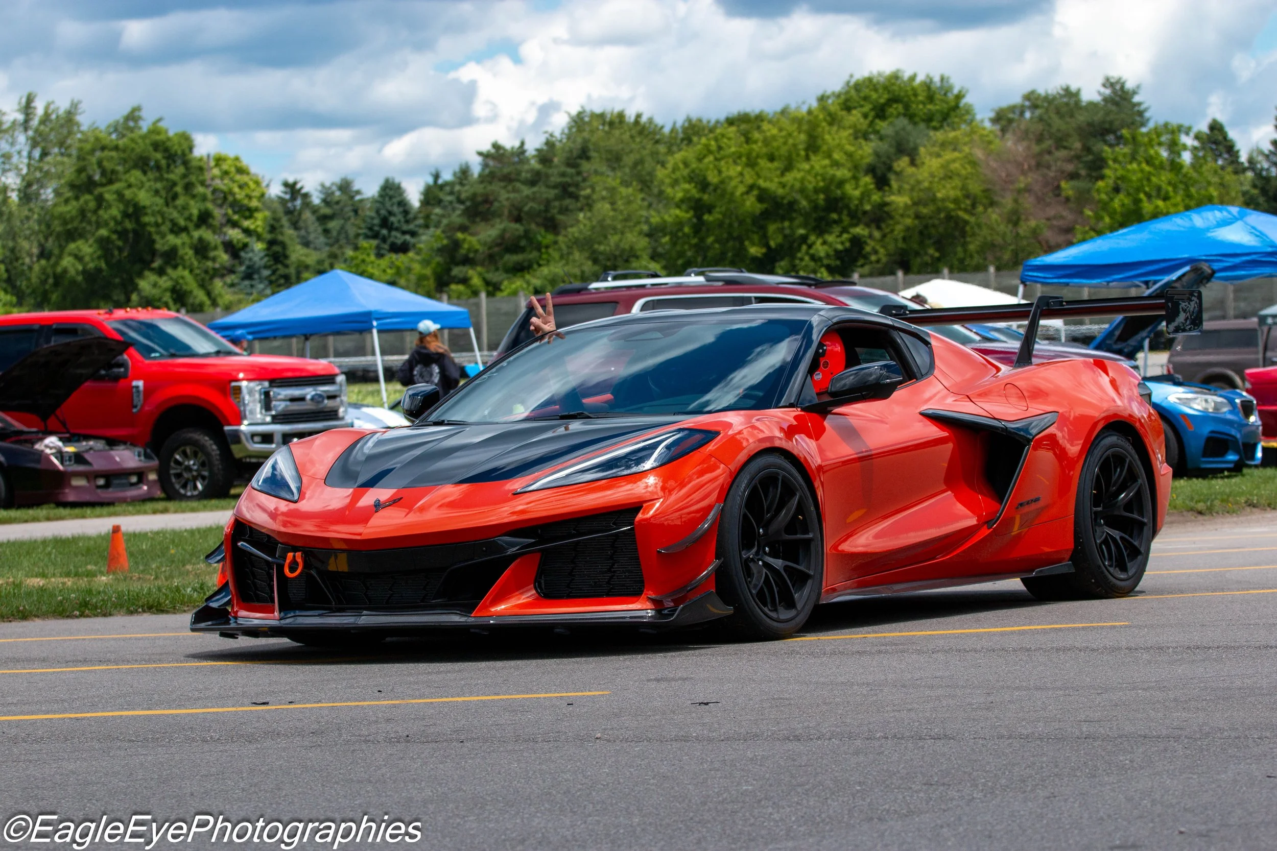 A red sports car driving on the road at an outdoor car event, with several parked cars, tents, and a person making a peace sign in the background, surrounded by green trees and cloudy sky.