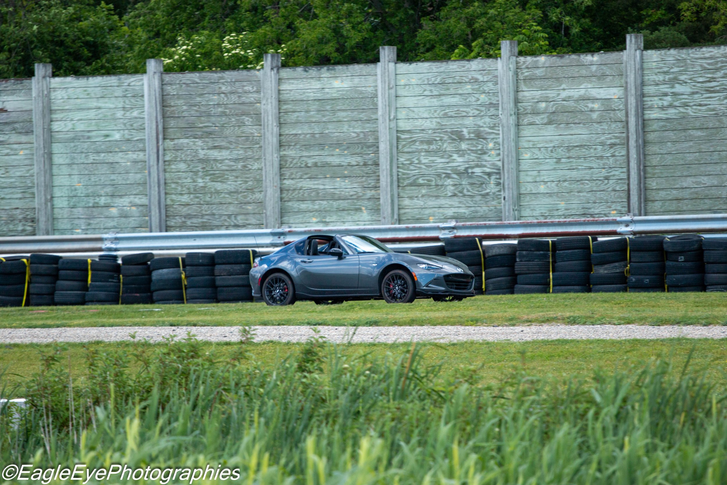 A gray sports car parked behind a barrier of black tires, with a tall wooden fence and green foliage in the background.