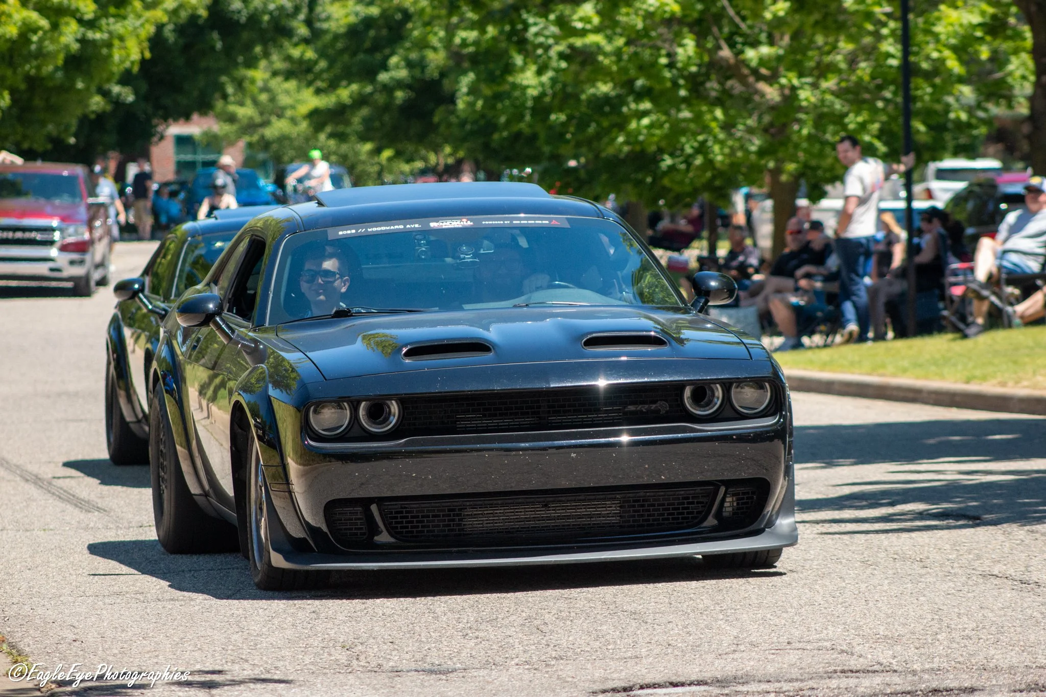 A black Dodge Challenger muscle car driving on a city street during a car show, with other cars and spectators in the background under trees.