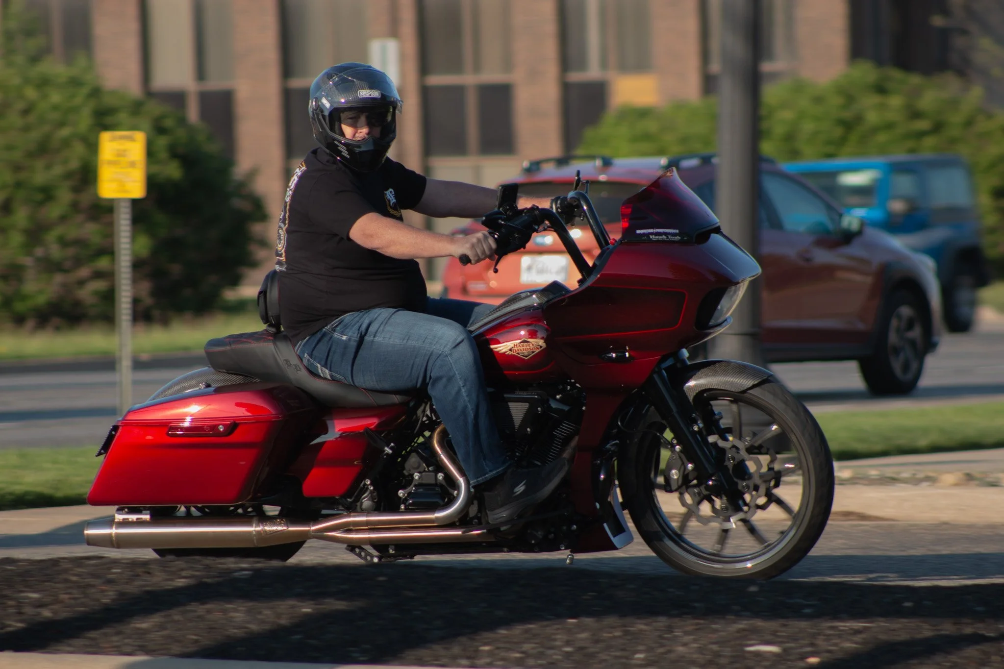 A man riding a red Harley Davidson motorcycle, wearing a black helmet and black shirt, on a city street with parked cars and a brick building in the background.