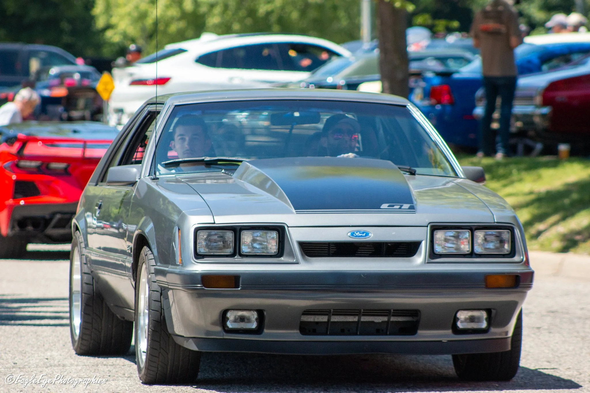 A silver classic Ford sports car being driven on a road with another vehicle behind it, with numerous parked cars and trees in the background, during a car show or gathering.