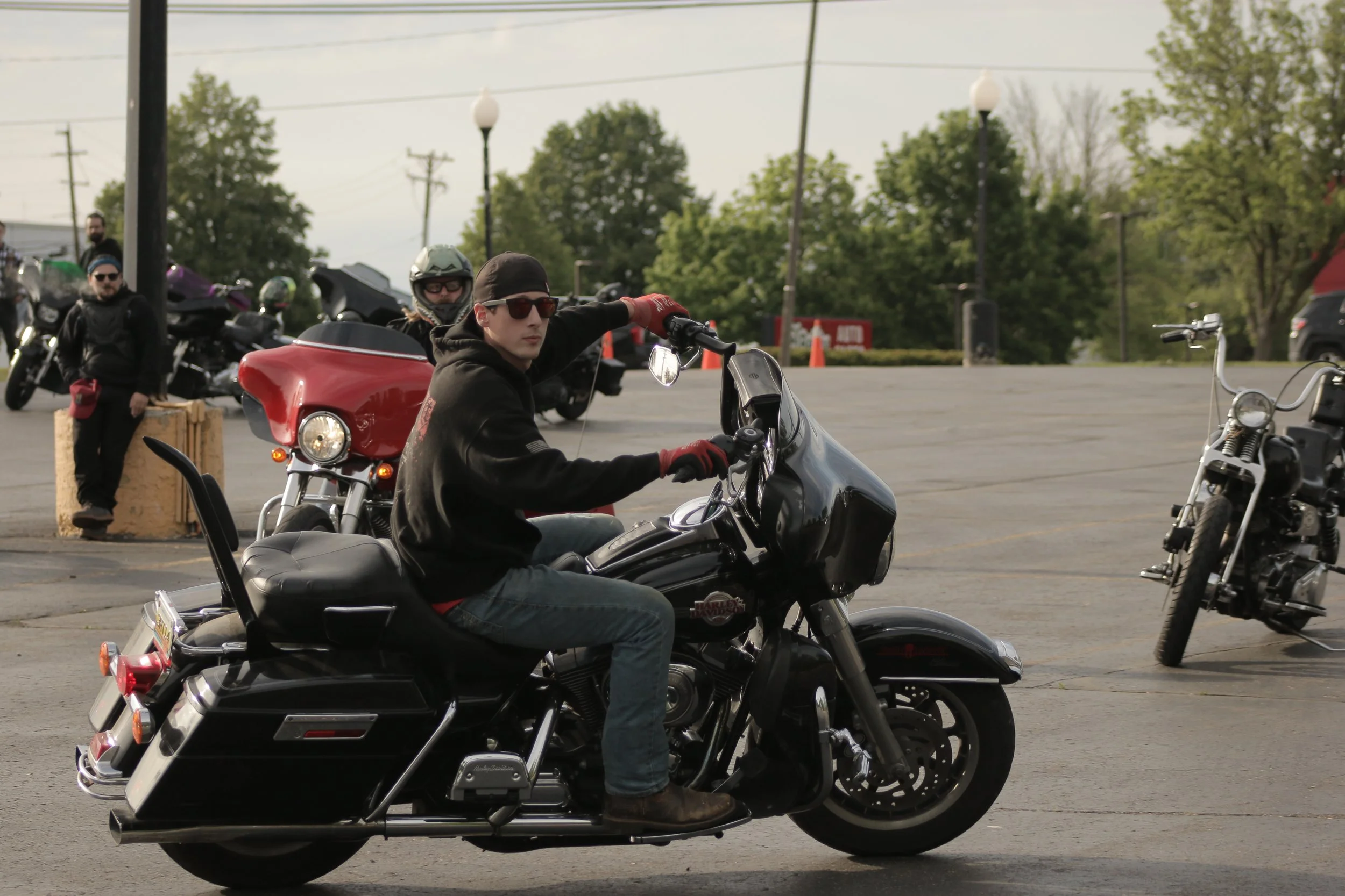 A man wearing sunglasses, a black hoodie, and red gloves riding a black motorcycle in a parking lot. Several motorcycles and people are visible in the background, with green trees and lamp posts surrounding the parking area.