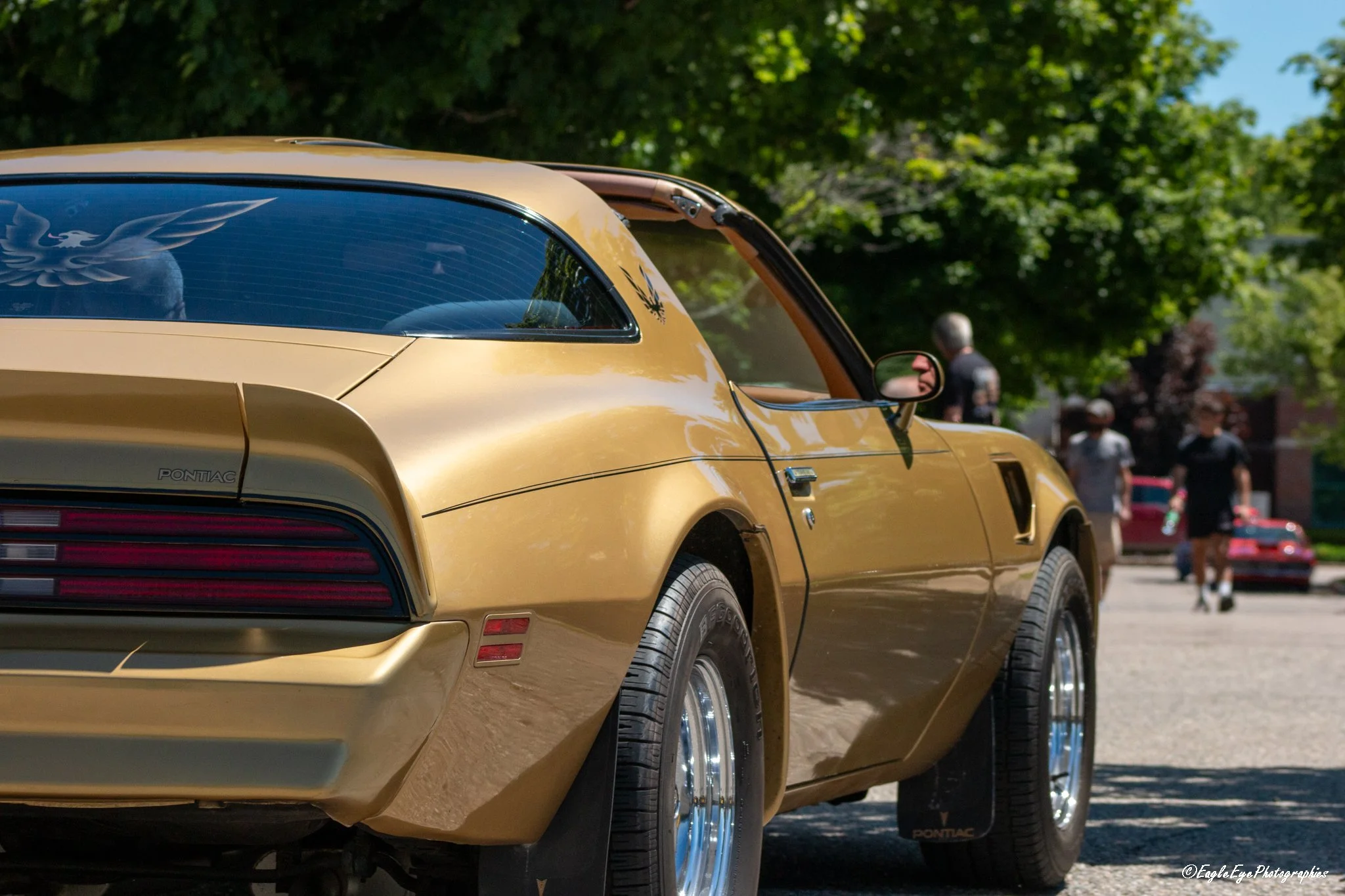 Close-up of a yellow Pontiac muscle car's rear side with a black Pontiac logo decal on the rear window, parked outdoors with trees and people in the background.