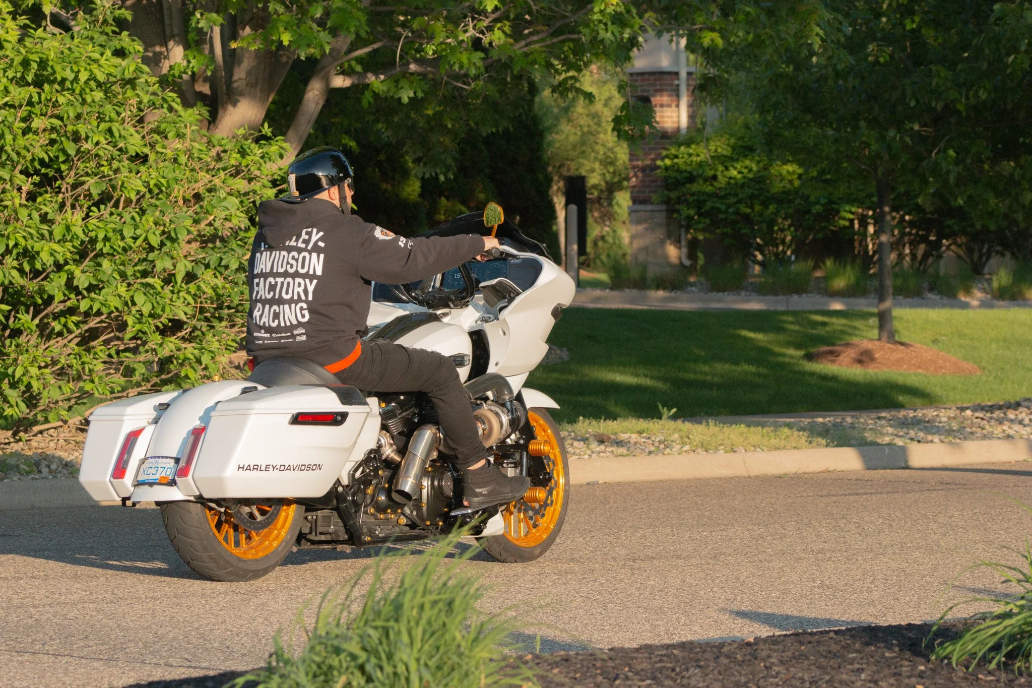A person wearing a Harley-Davidson hoodie and black helmet riding a white Harley-Davidson motorcycle with orange wheels along a suburban street lined with trees and houses.