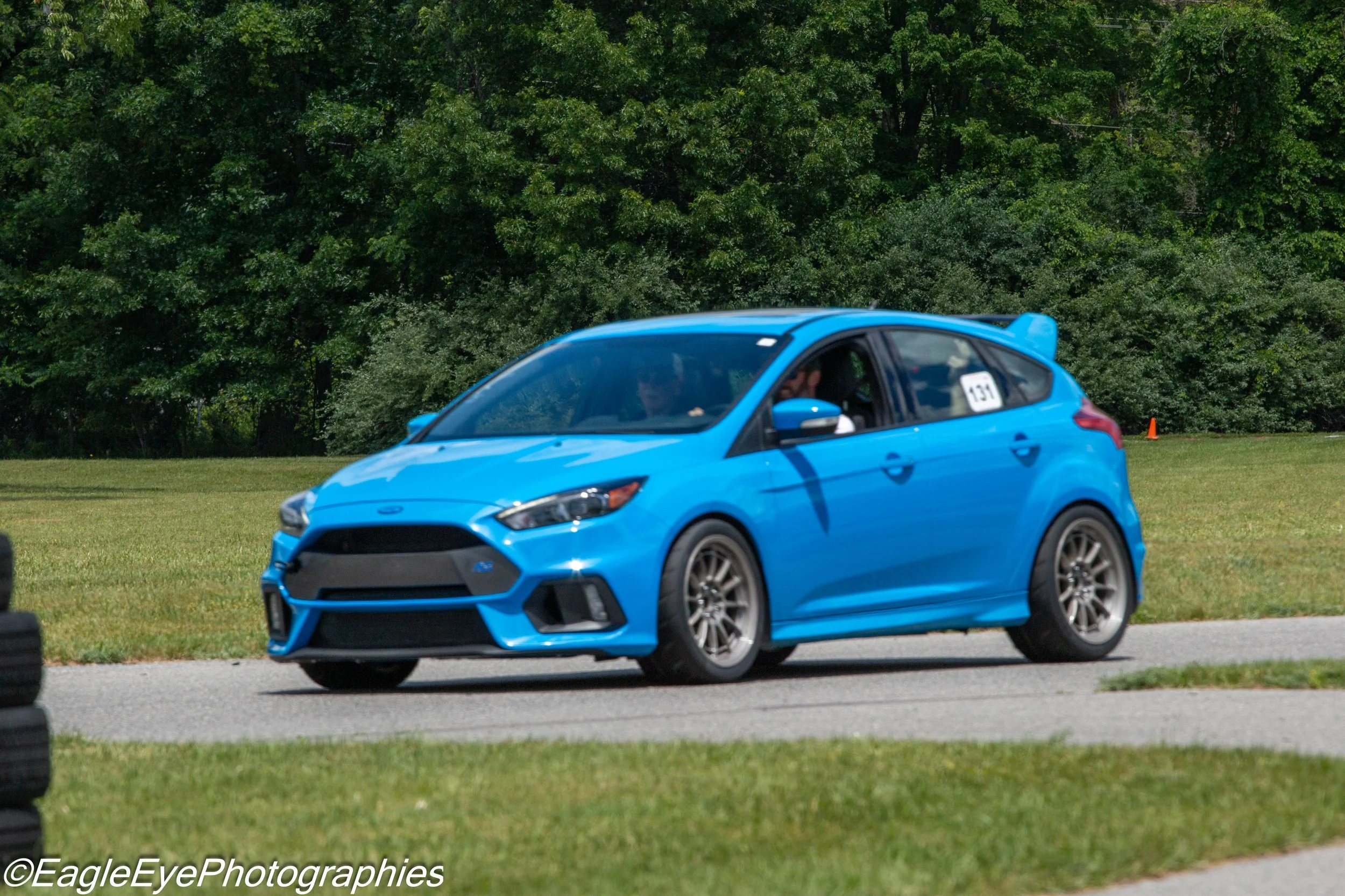 A blue compact car on a grassy field with trees in the background, part of a race or test event, with a small white number plate reading '131' on the side.