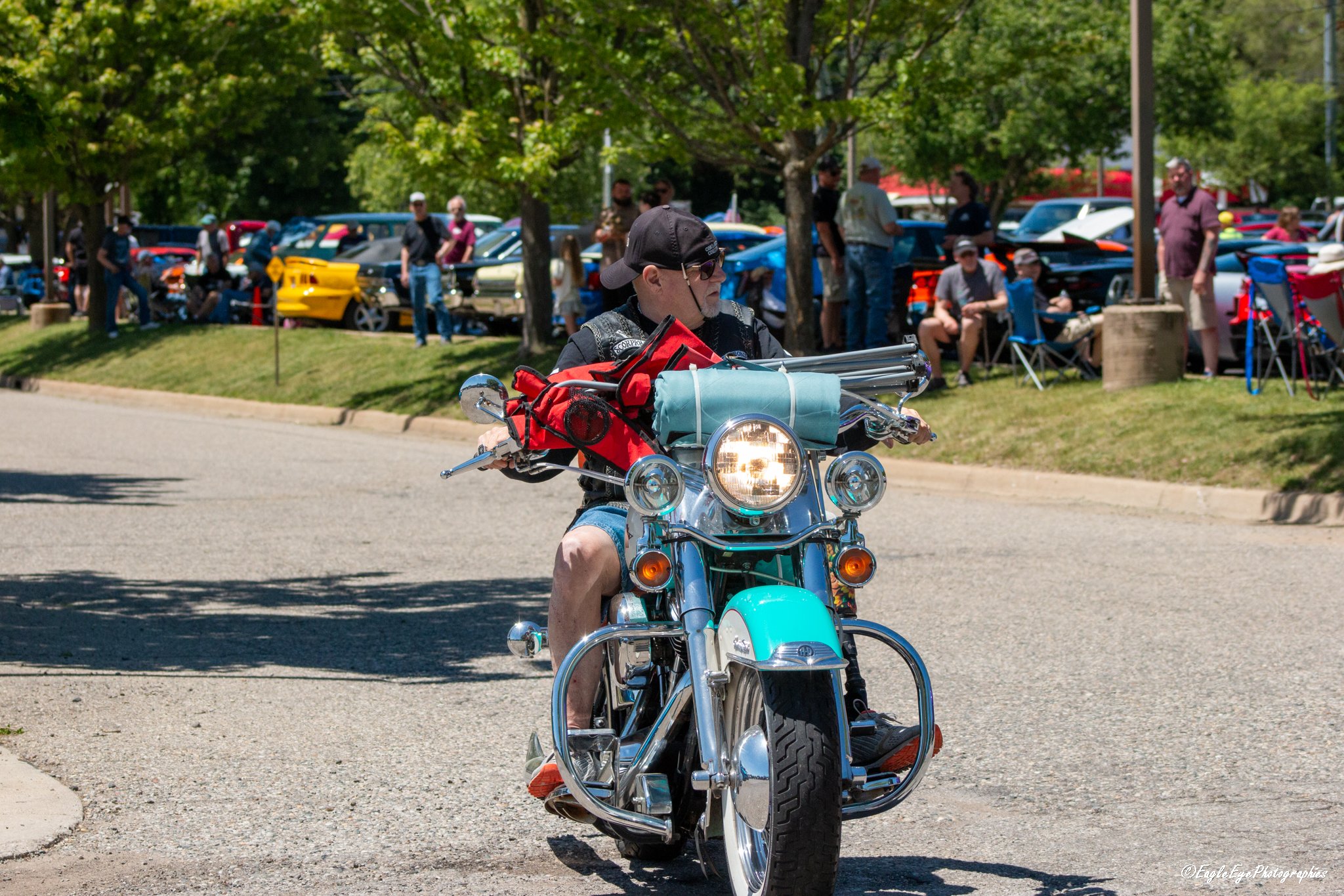 A man wearing a black cap and sunglasses riding a turquoise motorcycle at a car show, with classic and sports cars parked behind him under trees.