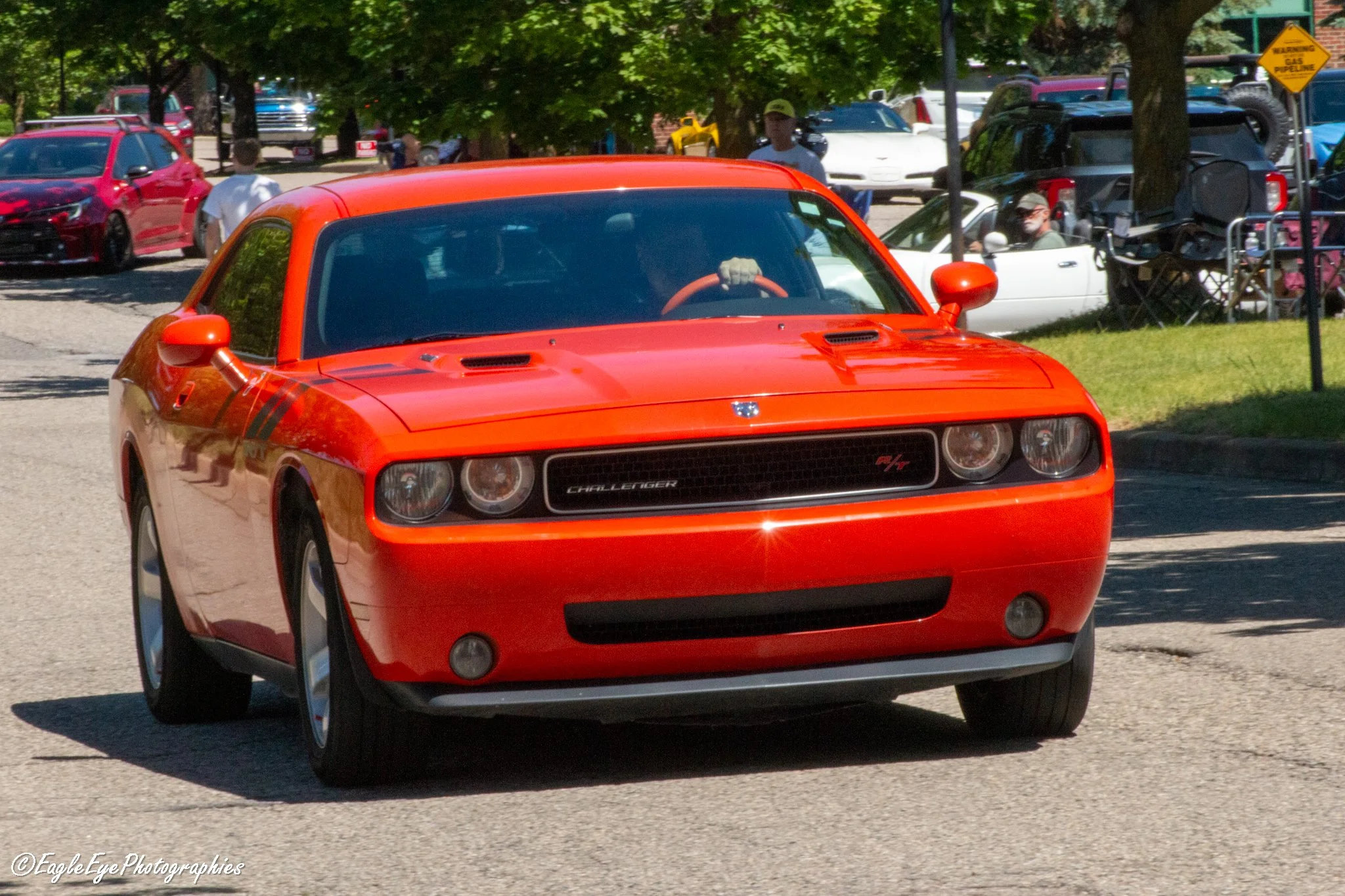 A red Dodge Challenger driving on a street with trees and parked cars in the background, including a white convertible and a black SUV.