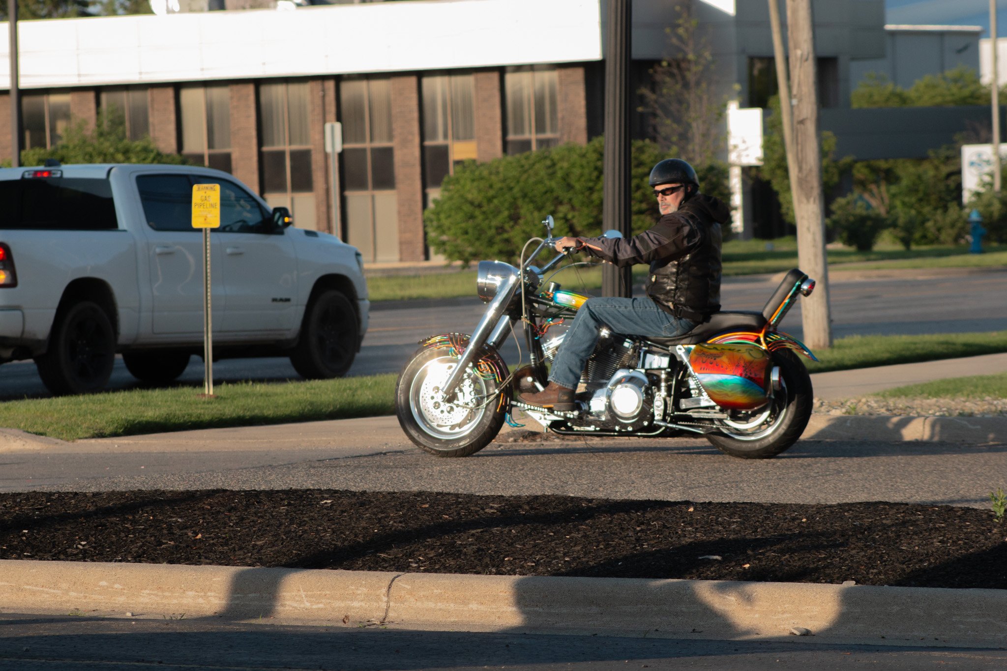 A man wearing a black helmet and sunglasses riding a colorful, rainbow-patterned motorcycle on the street. A white pickup truck is parked nearby, and a yellow 'Gas Pipeline' sign is visible. In the background, there are trees and a building with larg