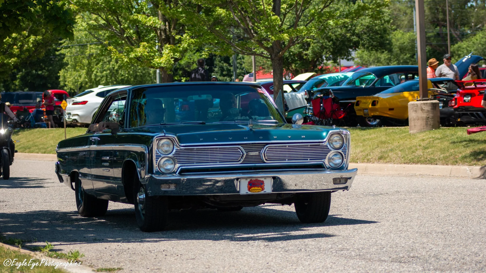 A vintage blue Plymouth car at a classic car show, parked in front of several other cars including a red and yellow sports car, with people walking around and looking at the vehicles under green trees.