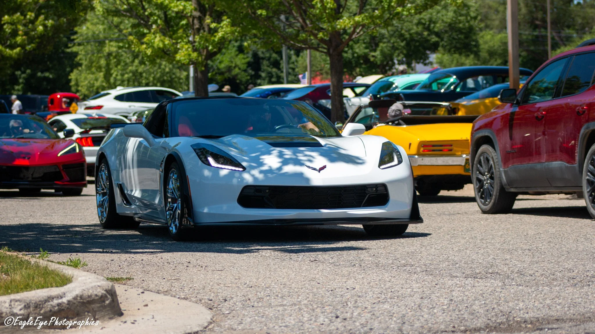 A white Chevrolet Corvette sports car parked among other cars at an outdoor car show, with trees and people in the background.