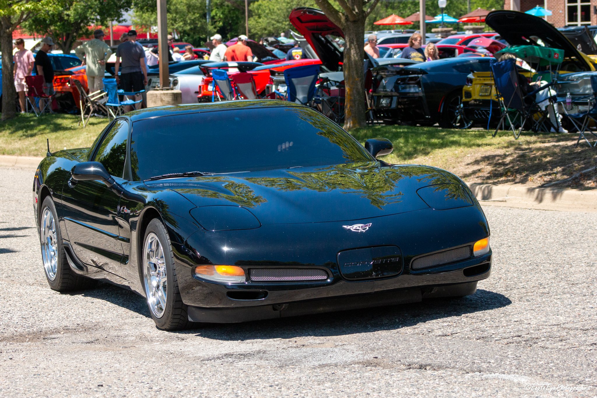 A shiny black sports car parked outdoors at a car show with its hood closed, in front of a crowd of people and many other cars parked in the background.