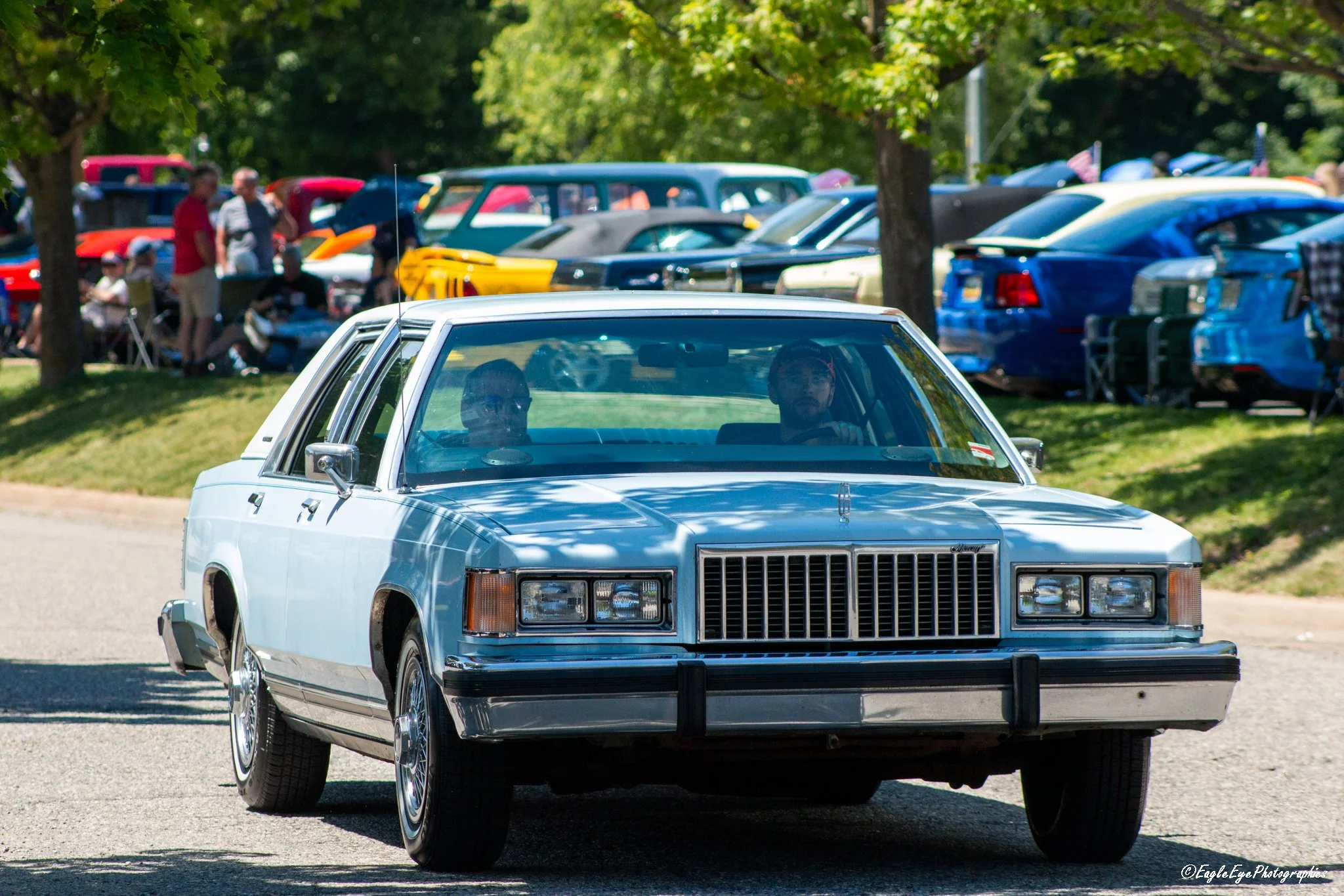 A silver vintage car with people inside driving past a crowded outdoor car show with numerous classic cars parked under trees.