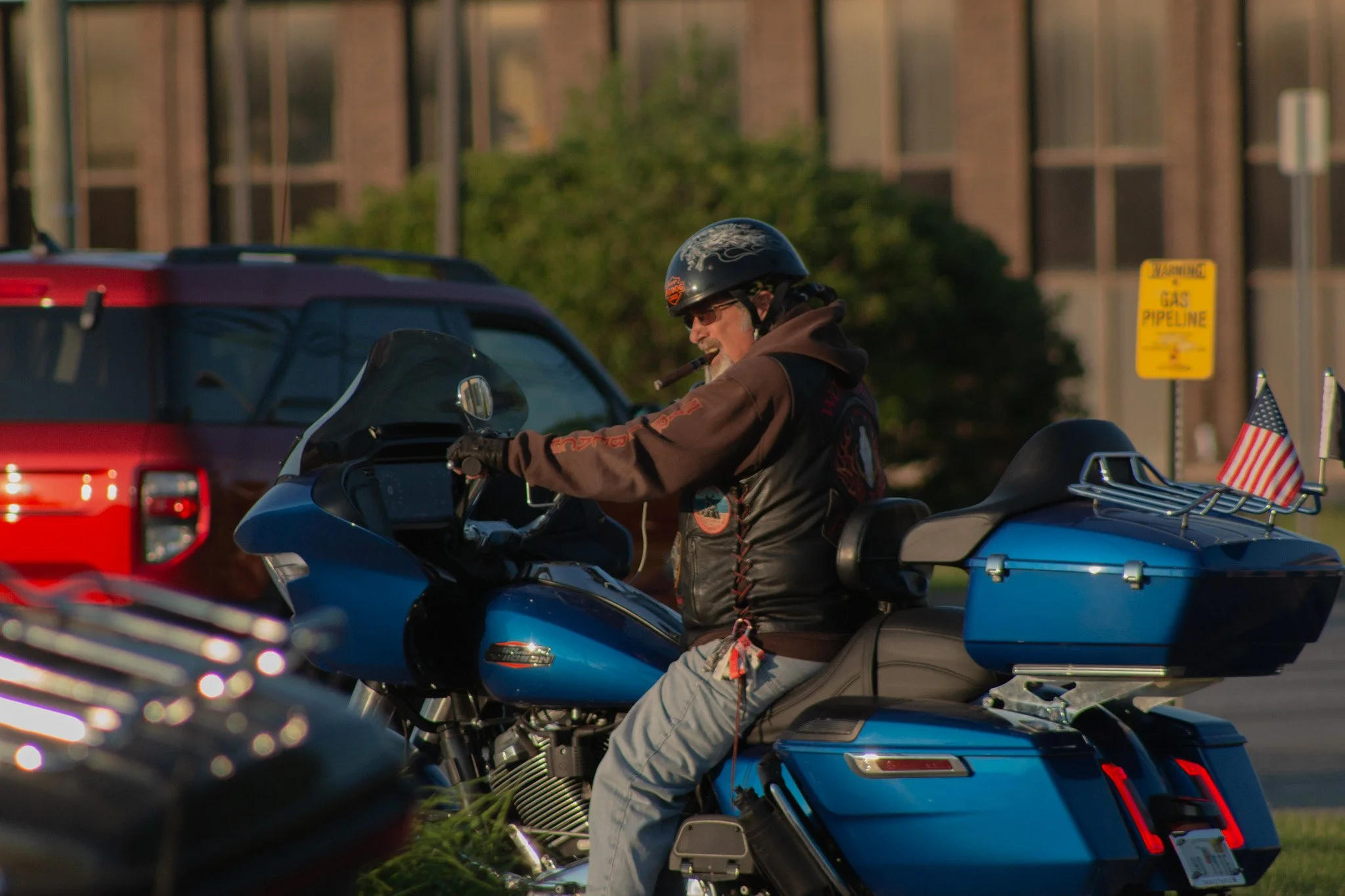 A man wearing a black helmet, sunglasses, sweatshirt, and vest riding a blue motorcycle with an American flag on the luggage compartment. There is a red vehicle and a yellow gas pipeline warning sign in the background.