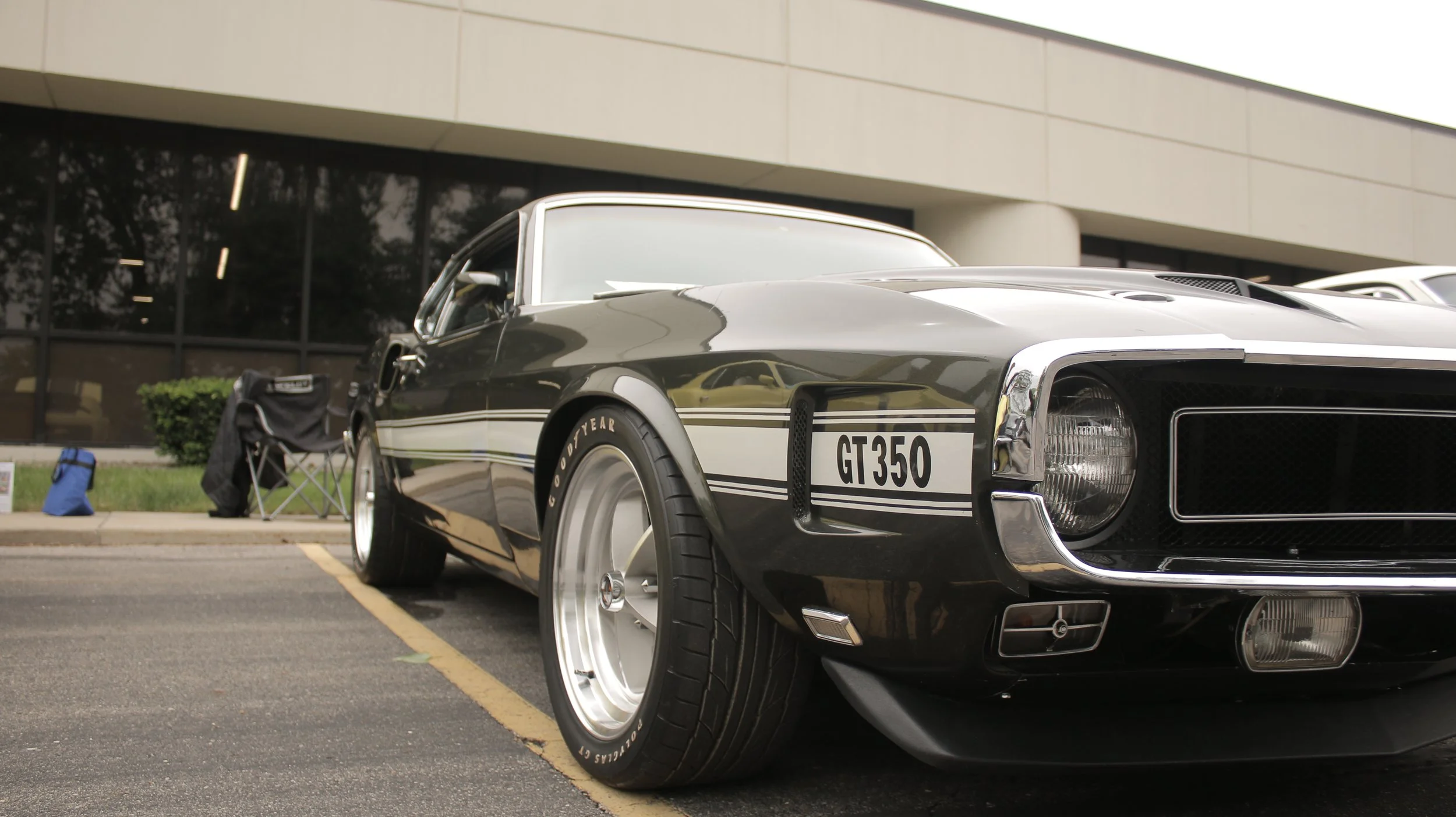 A vintage black car with white racing stripes and a GT350 emblem stationed in a parking lot near a modern office building, with a camping chair, cooler, and small bag visible nearby.