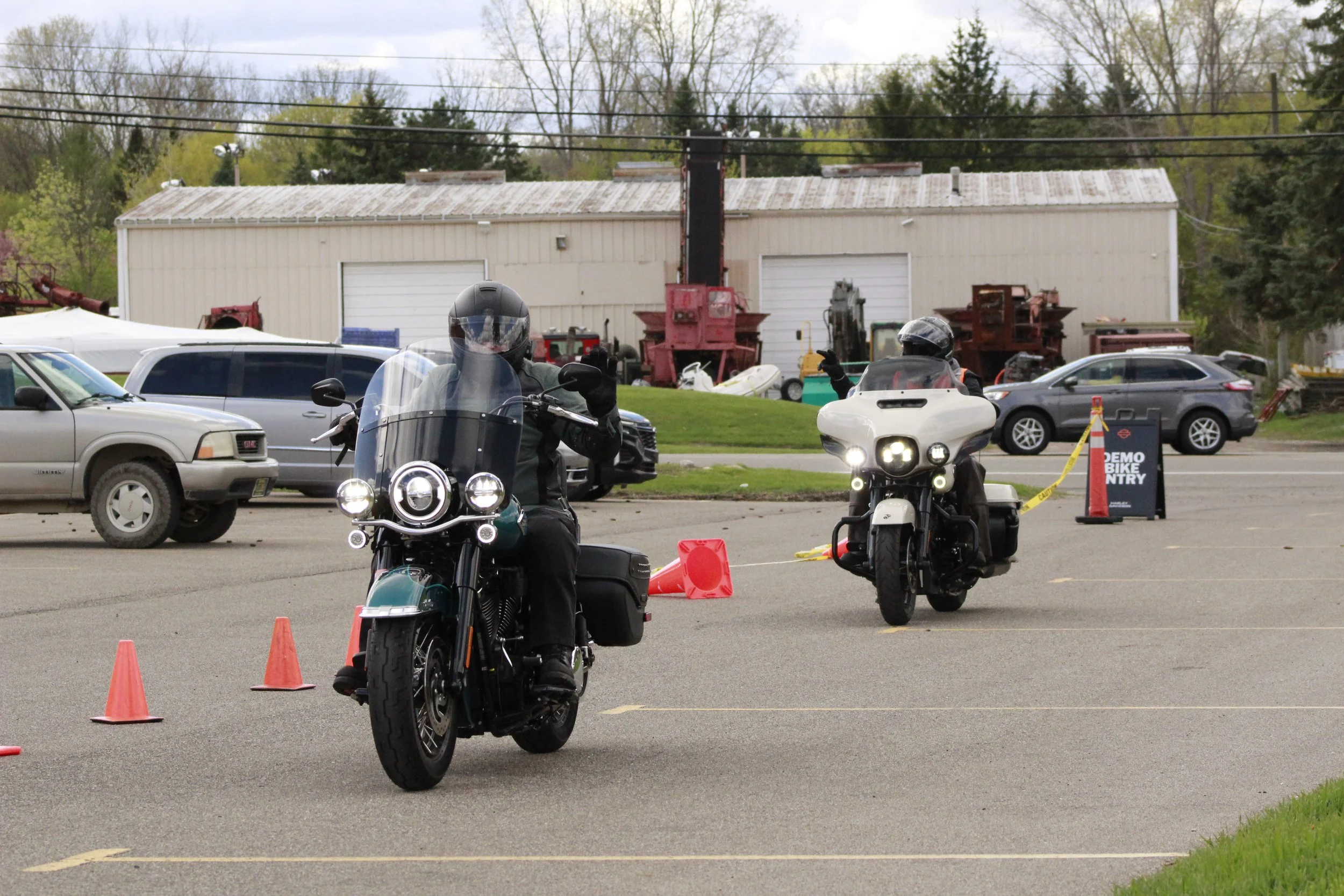 Two motorcyclists wearing helmets and riding gear riding side-by-side on a street. Orange cones mark a designated area. Behind them is a parking lot with parked cars and a building with closed garage doors. Trees and power lines are visible in the ba