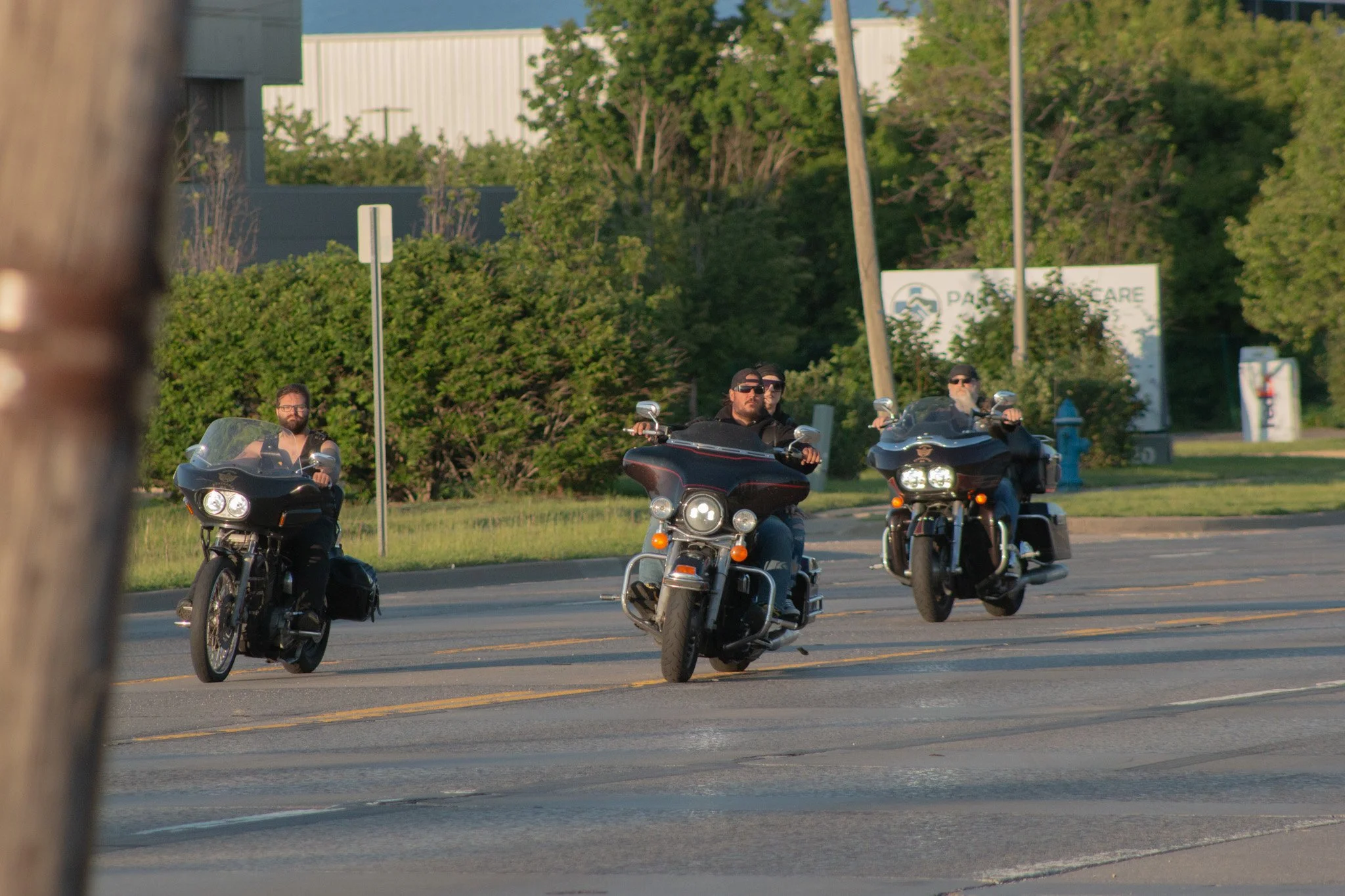 Four men riding motorcycles on a street, with trees and a parking lot in the background.