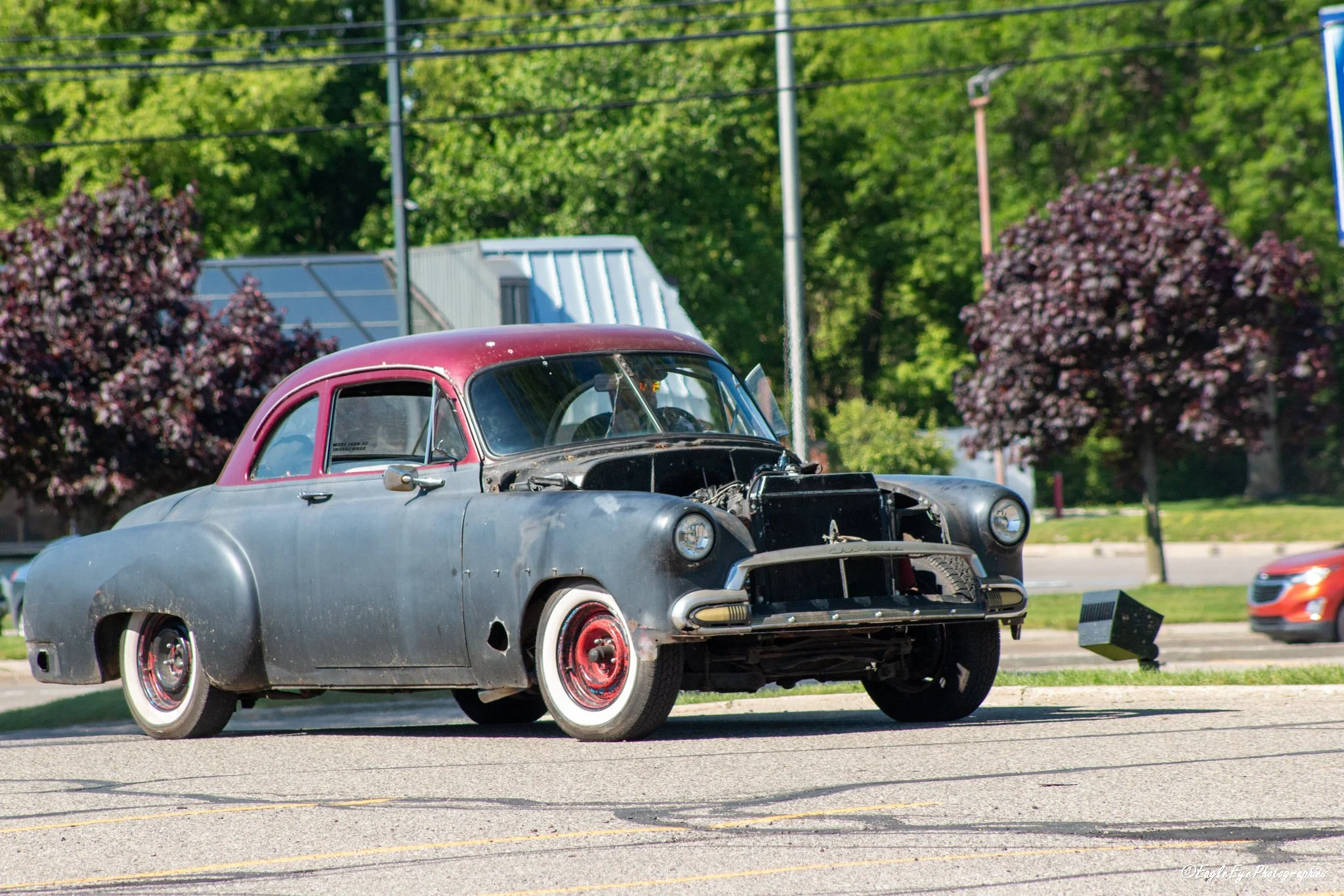 Two vintage cars on display outdoors with trees and power lines in the background. The cars are old models, one metallic gray and the other with a faded red roof, with the front bumper and grill of one car visible.