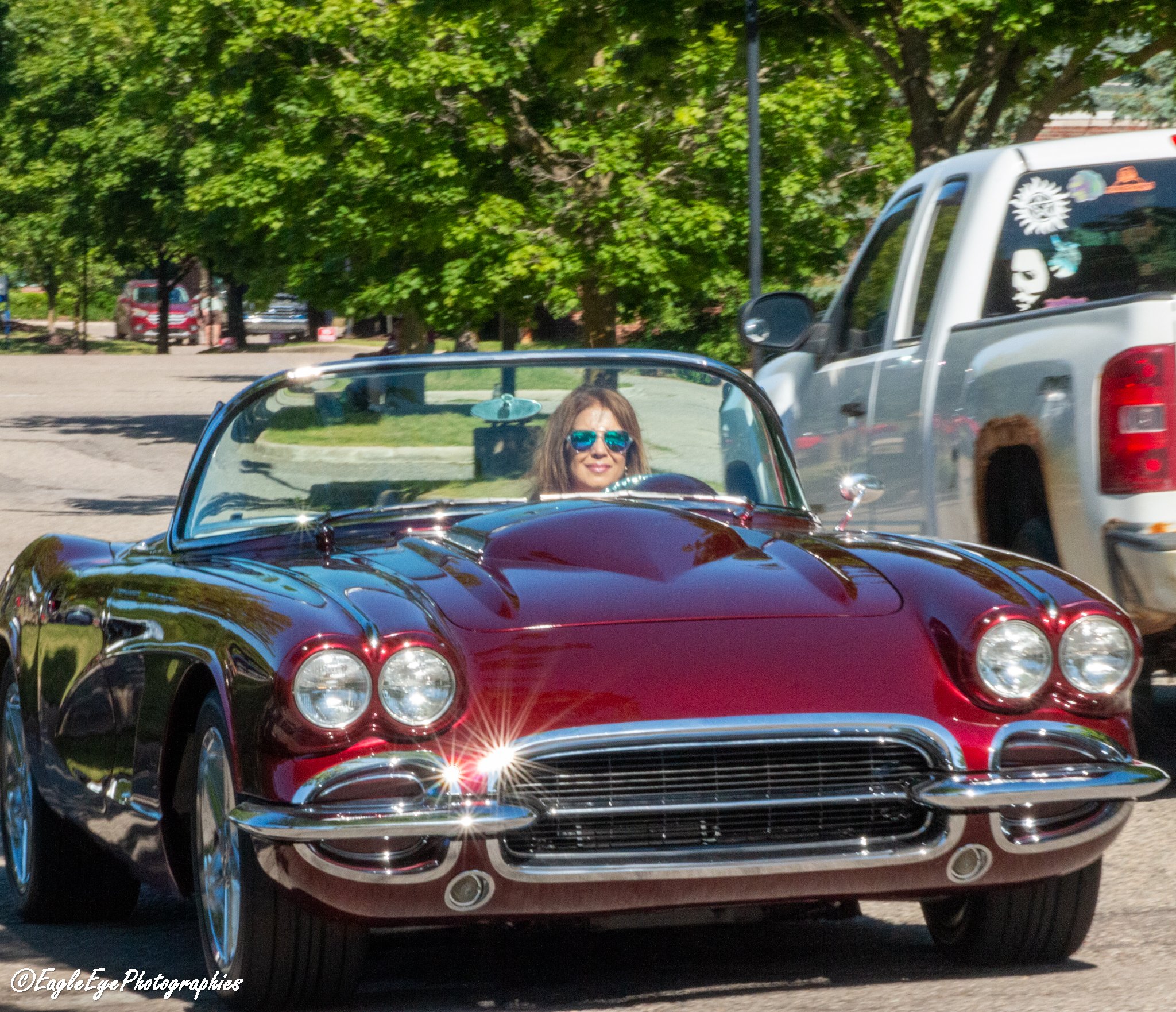 A woman with blue sunglasses smiling in a red vintage convertible car, with a parking lot, trees, and other vehicles in the background.