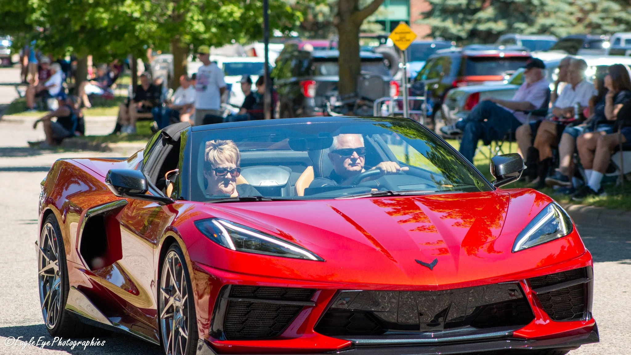 A red Corvette sports car driving through a park with people sitting on benches under trees in the background.