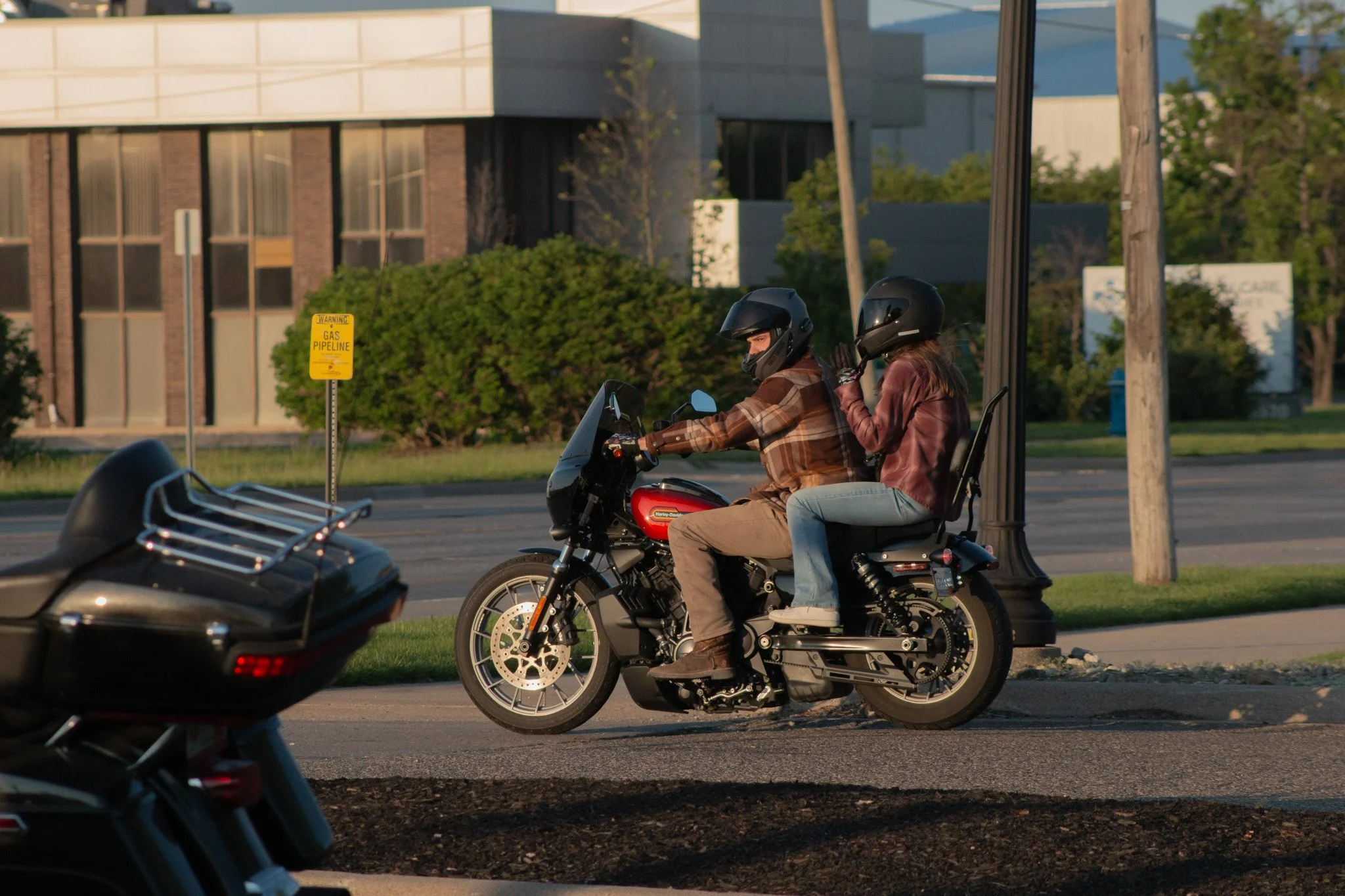 A man and woman wearing helmets, riding a motorcycle together on a city street. The man is driving, and the woman is sitting behind him, holding onto his waist. The background shows a modern building, green bushes, a utility pole, and a sign that say