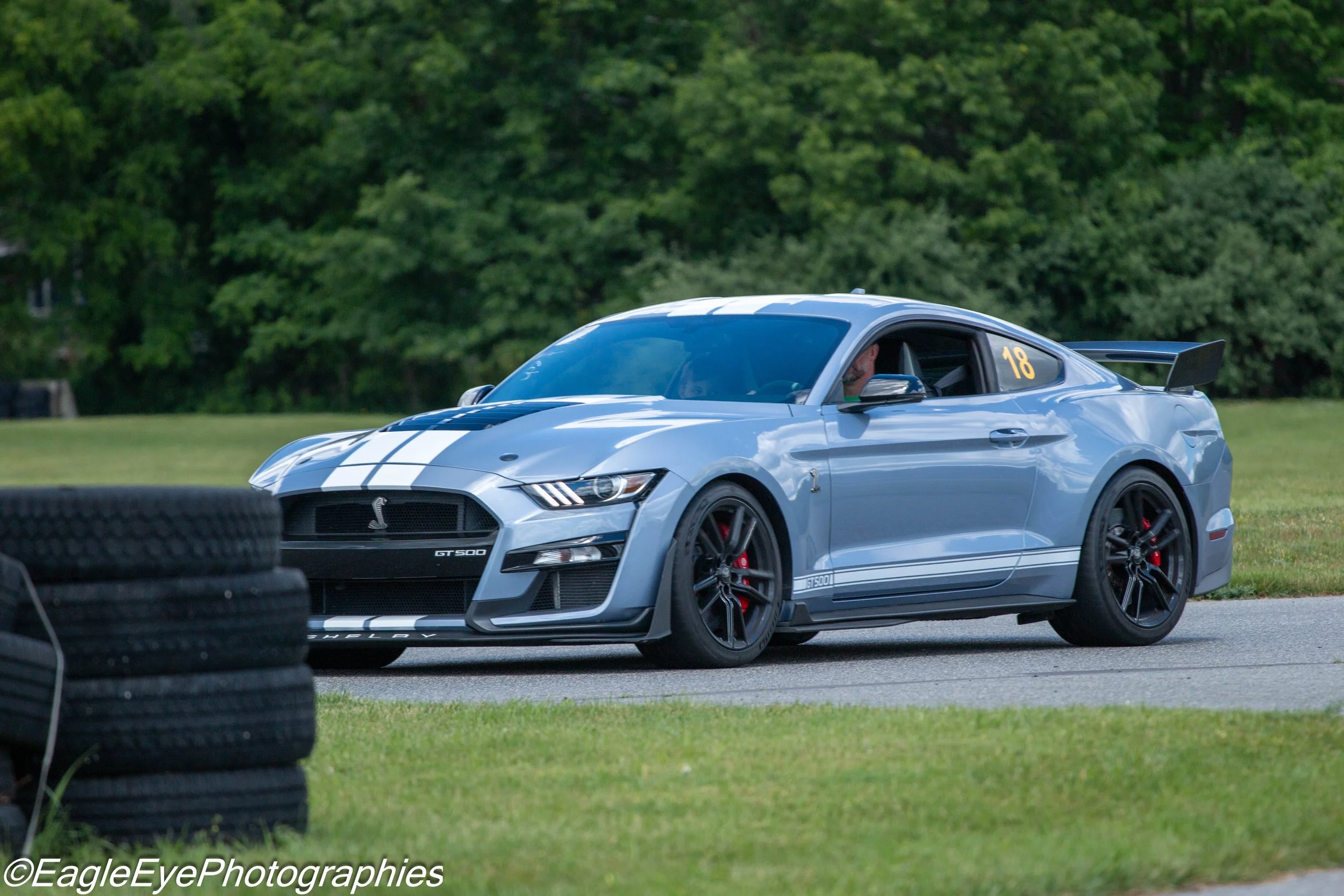 A light blue Ford Mustang Shelby GT500 race car with the number 18 on the window, driving on a race track; tires in the foreground, trees in the background.