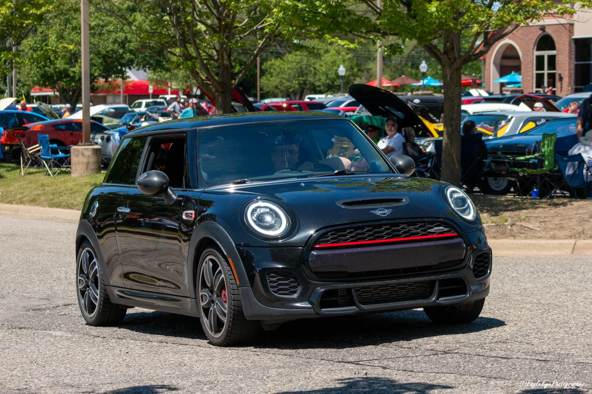 A black Mini Cooper parked in front of a busy outdoor car show with numerous cars and people in the background, under trees.