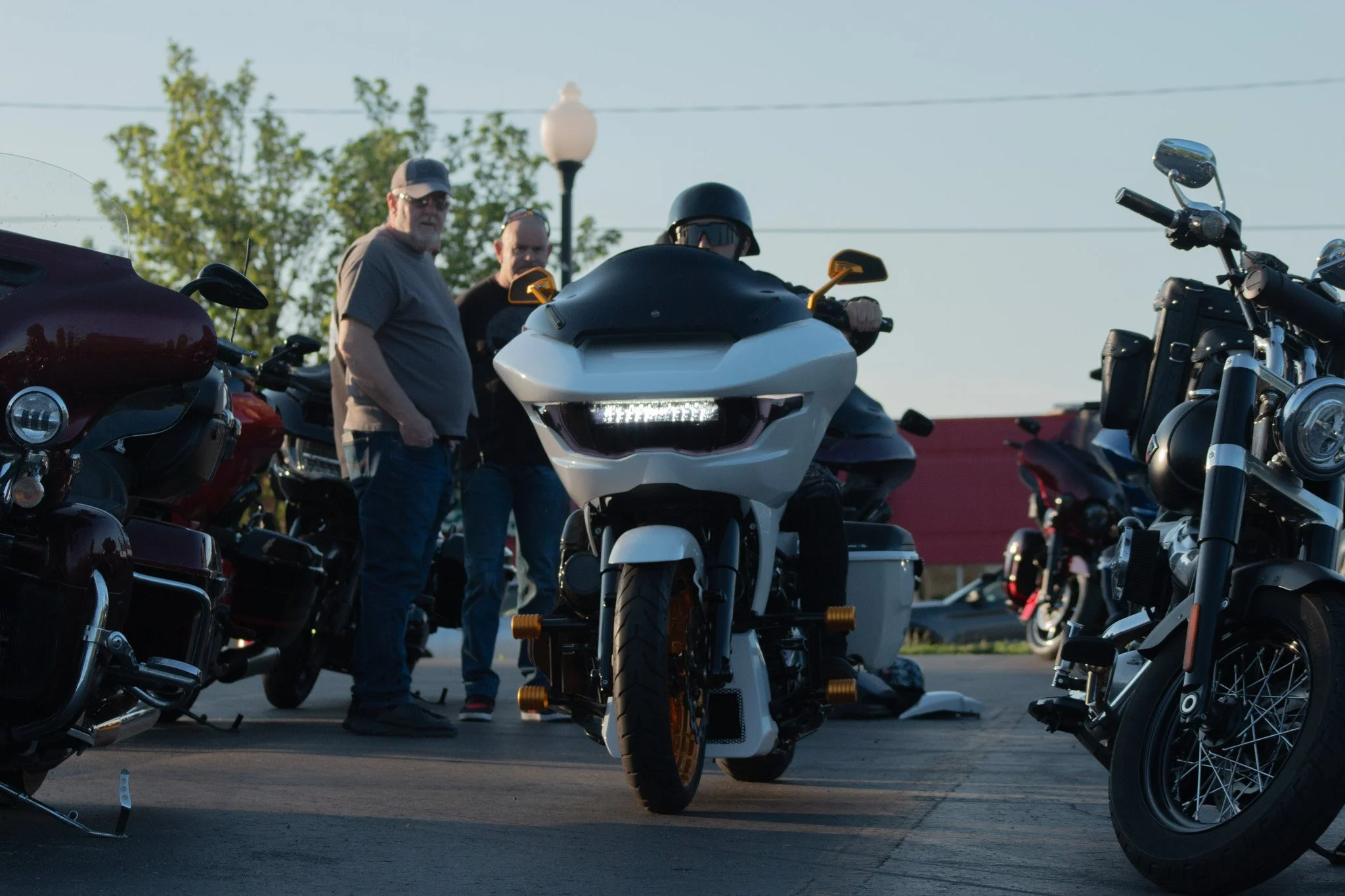 Several men are gathered around a collection of motorcycles and a white touring trike motorcycle, in an outdoor parking lot with greenery and overhead power lines in the background.