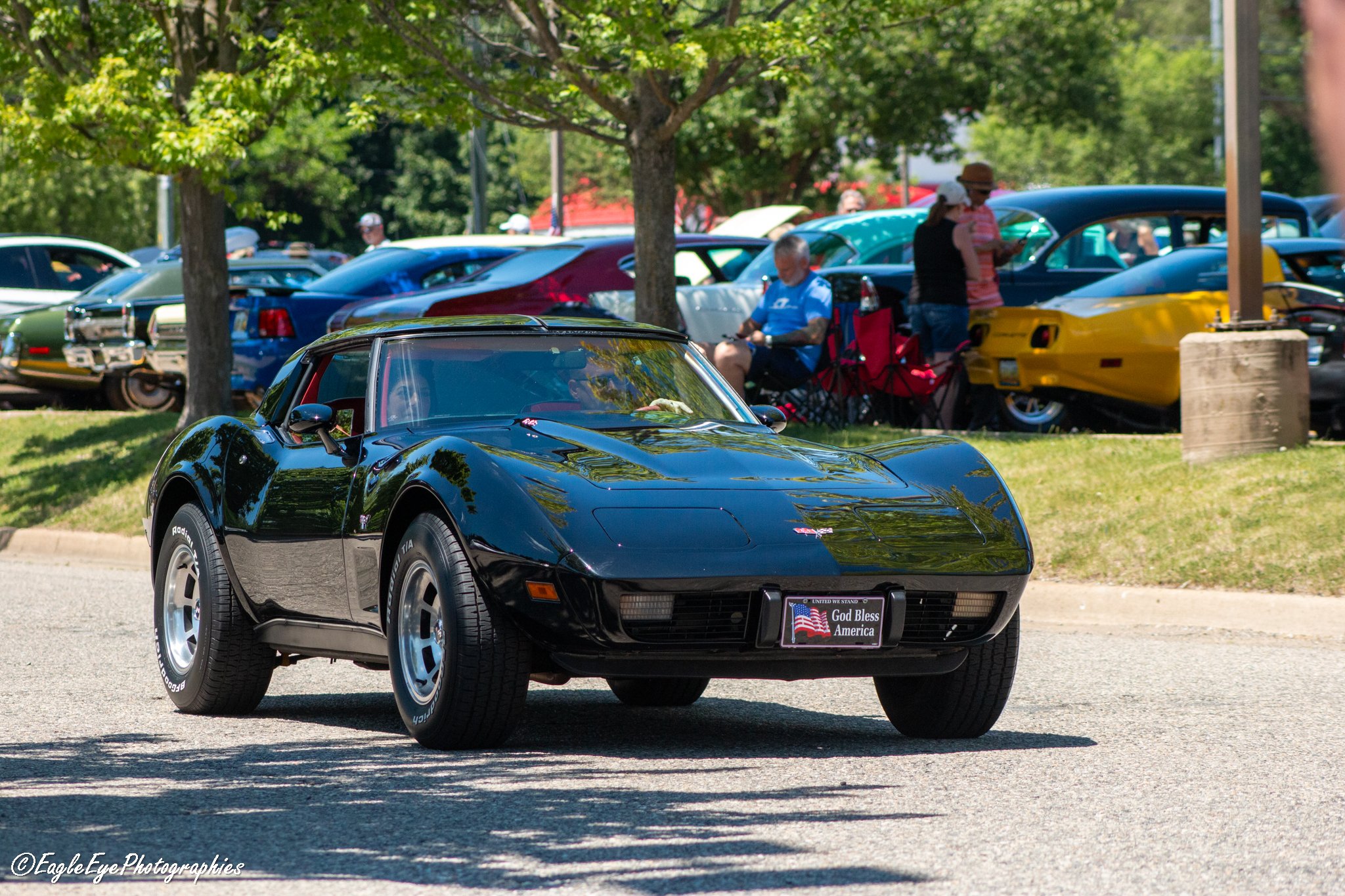 A black Chevrolet Corvette sports car parked outdoors with a patriotic license plate that reads 'God Bless America' and an American flag on it. The background features a car show with numerous vintage and classic cars, trees, and people walking and t