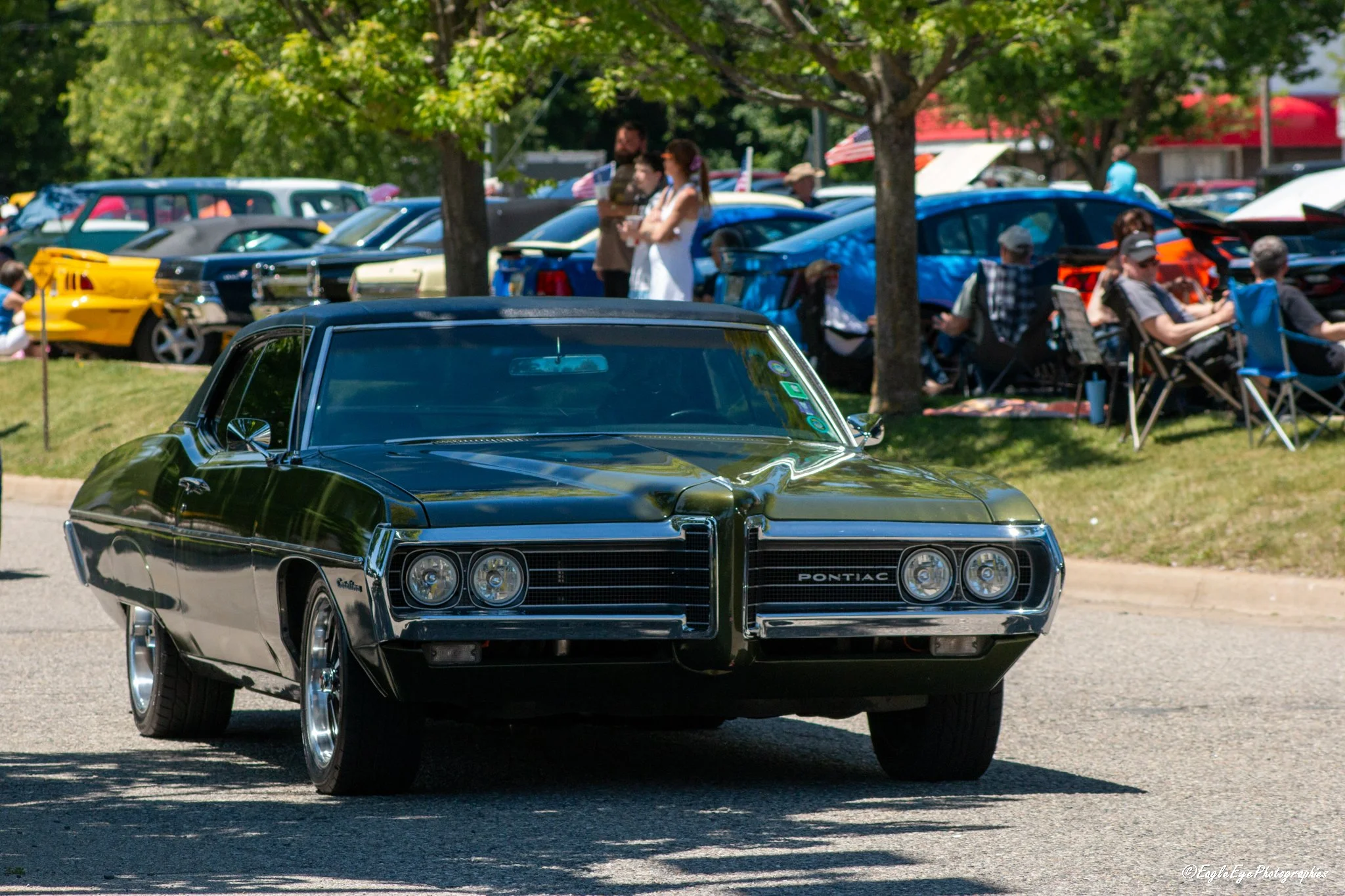 A black vintage Pontiac car driving in a car show with a crowd of people and multiple parked cars in the background.