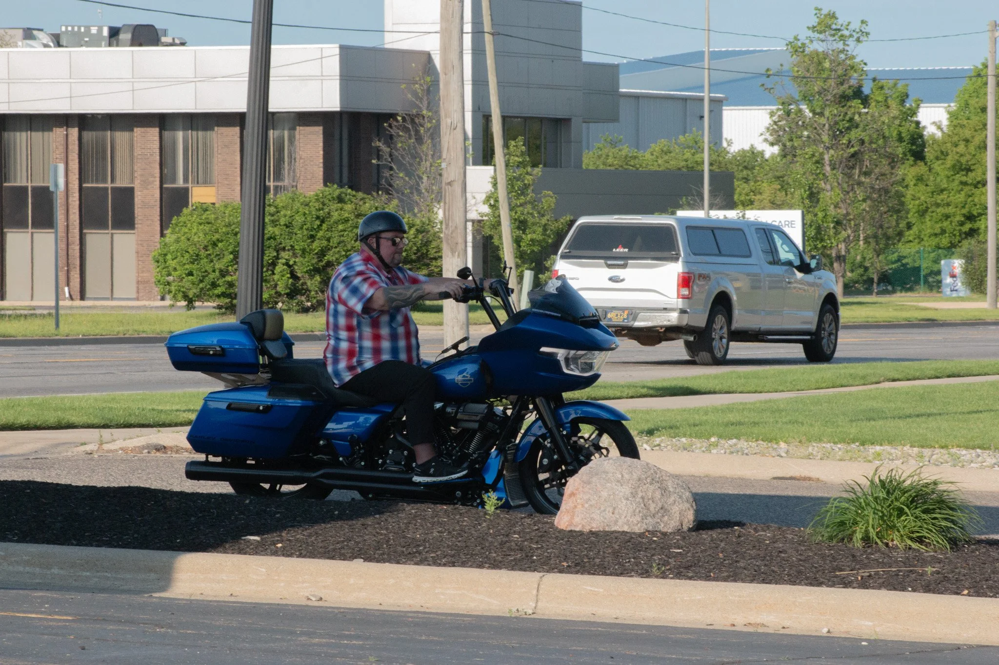 A man wearing sunglasses, a black helmet, and a red, white, and blue plaid shirt riding a blue motorcycle on a city street. There are cars and buildings in the background, along with trees and utility poles.