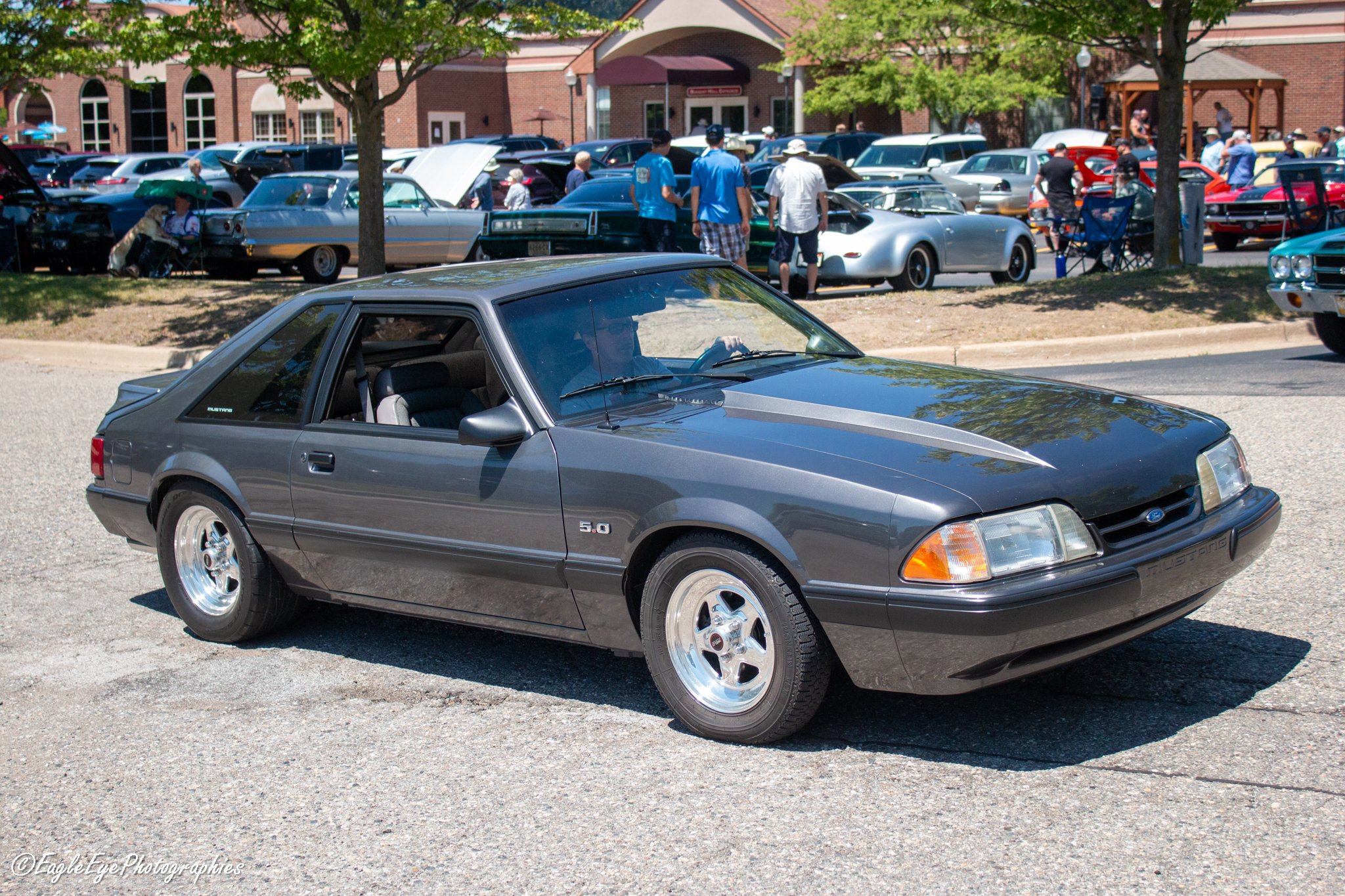 A classic gray Ford Mustang with a black hood parked in the foreground at a car show, with vintage cars and people gathering in the background under trees and a brick building.