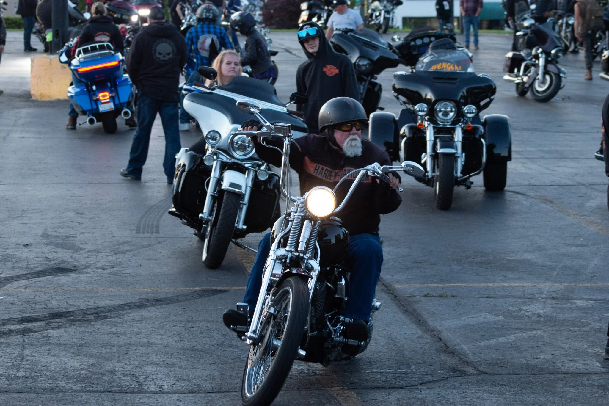 A man with a beard riding a black motorcycle, wearing a black helmet and sunglasses, in front of a group of motorcyclists and spectators at an outdoor gathering.