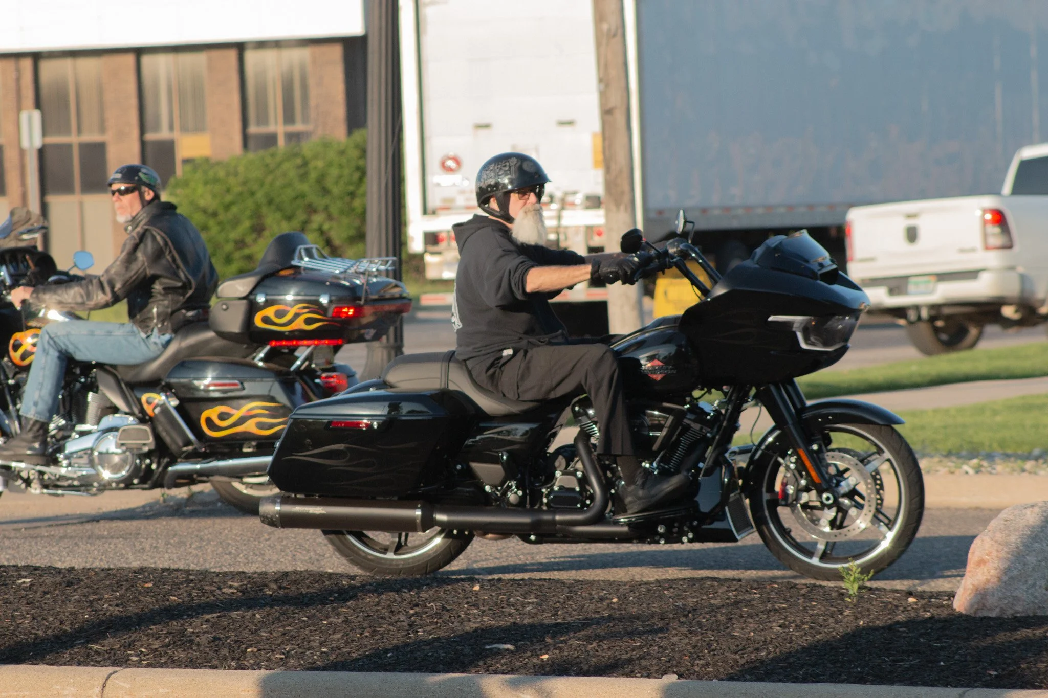 Two men riding black motorcycles on a street, both wearing helmets and dark clothing, with trucks and buildings in the background.