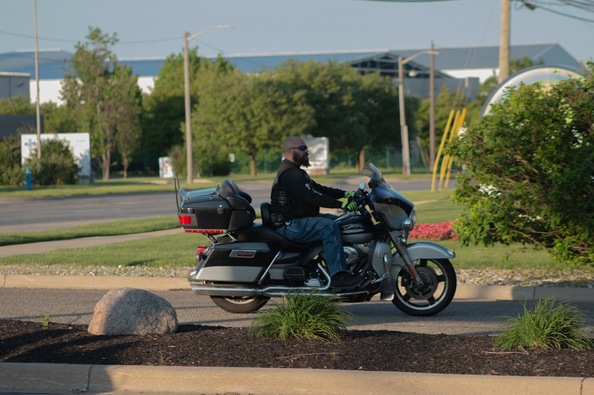 A man with a beard, wearing sunglasses, a black jacket, and gloves, is riding a black and gray touring motorcycle along a suburban street during daytime. There are trees, power lines, and buildings in the background, with a green shrub and flowers in