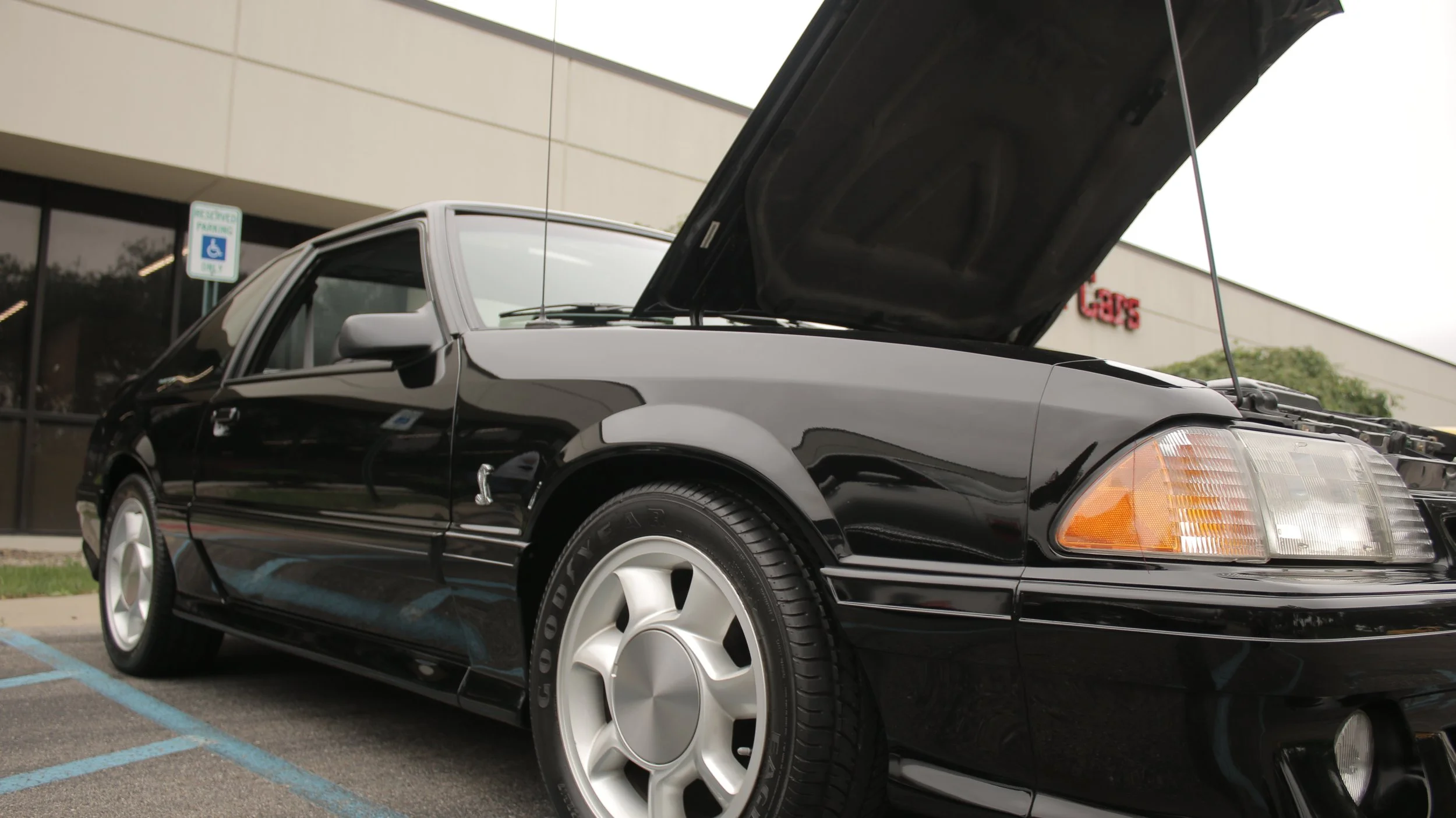 A black sports car with its hood open parked in a parking lot near a building with a sign that reads 'GAPS' and a visible handicap parking sign.