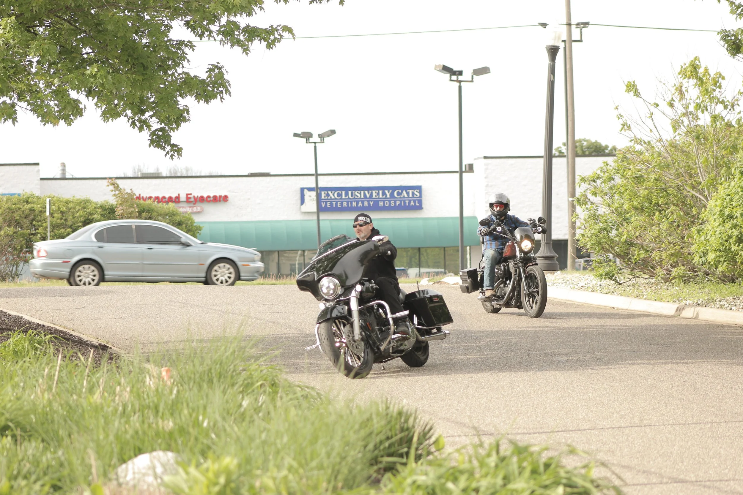 Two motorcyclists riding on the street near a veterinary hospital, with a tree and a silver car passing by in the background.