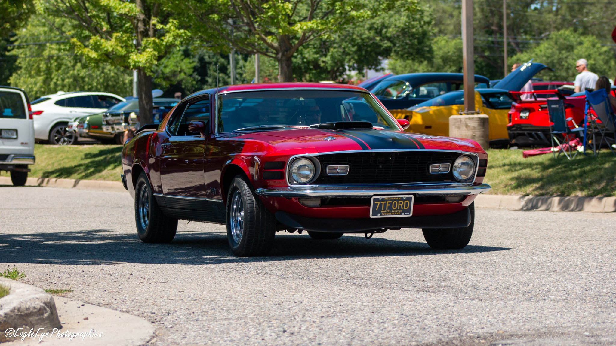 Red vintage Ford Mustang with black racing stripes parked at a car show, with other cars and people in the background under trees.
