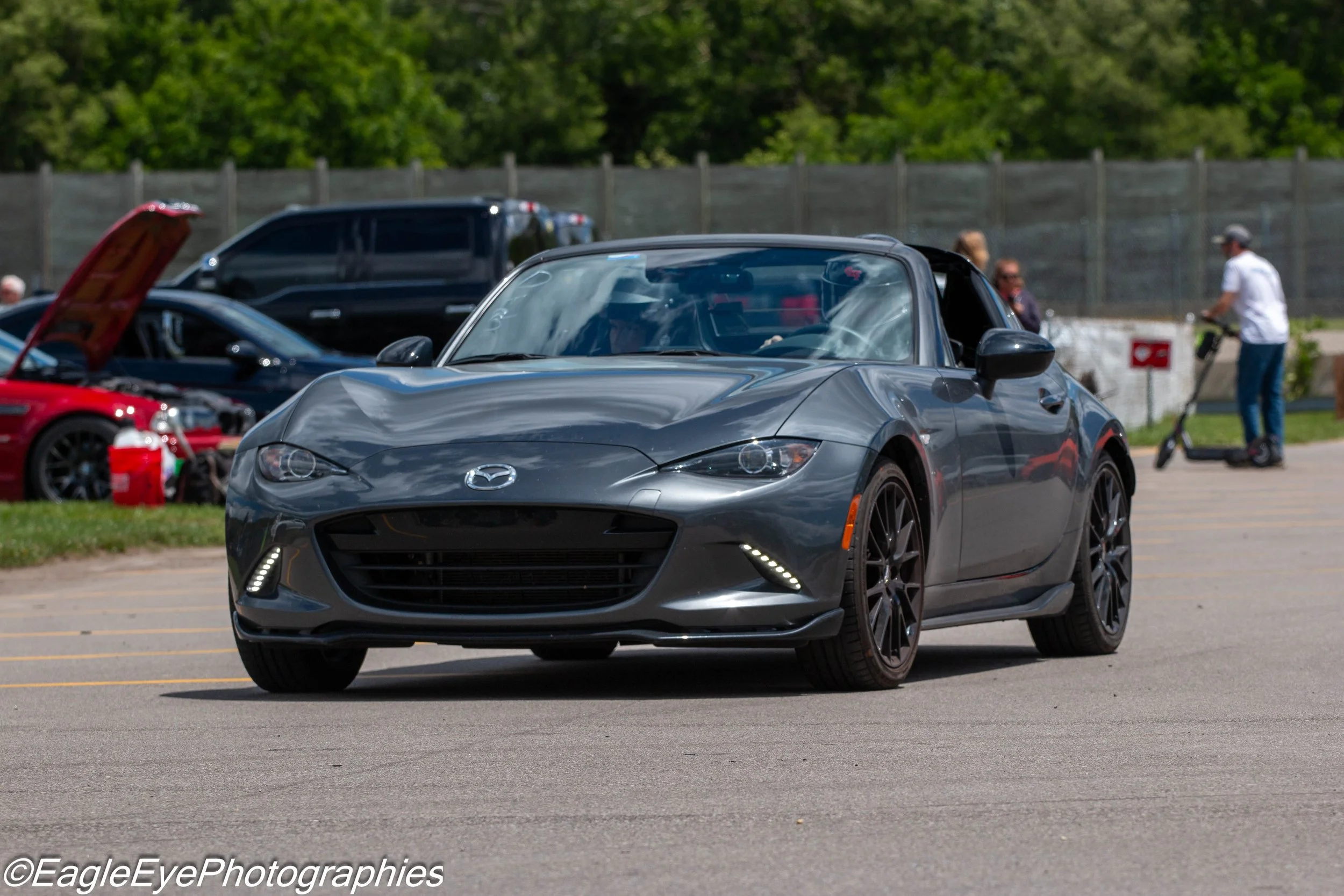 A gray Mazda MX-5 Miata sports car driving in a parking lot during daytime, with other cars and people in the background.
