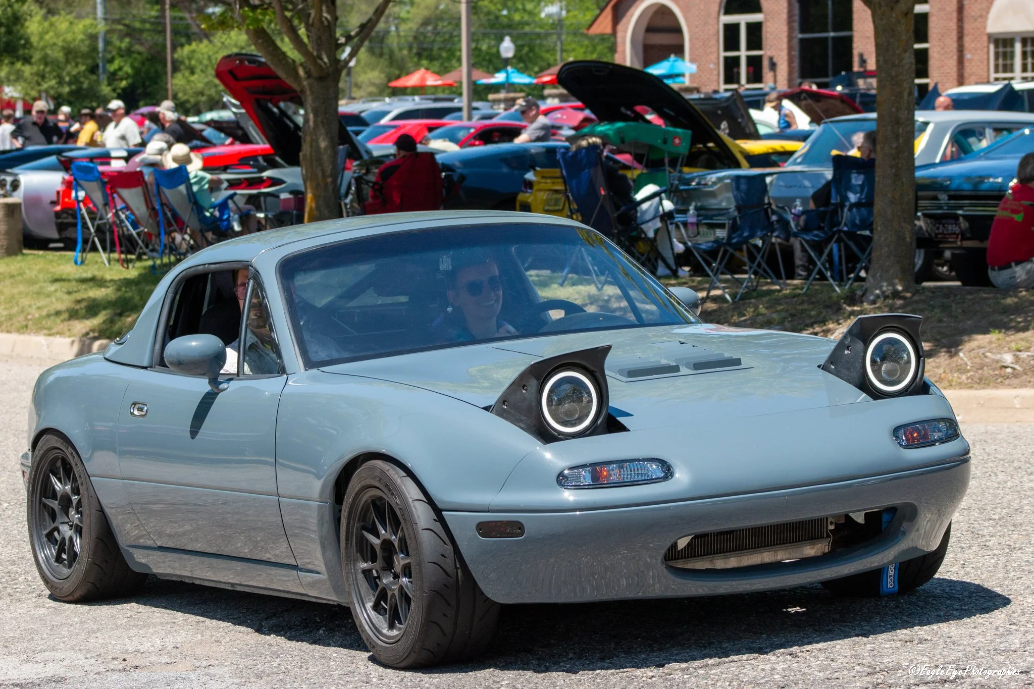 A silver compact sports car with custom black wheels and headlights parked outdoors during a car show, with many other cars and people in the background.