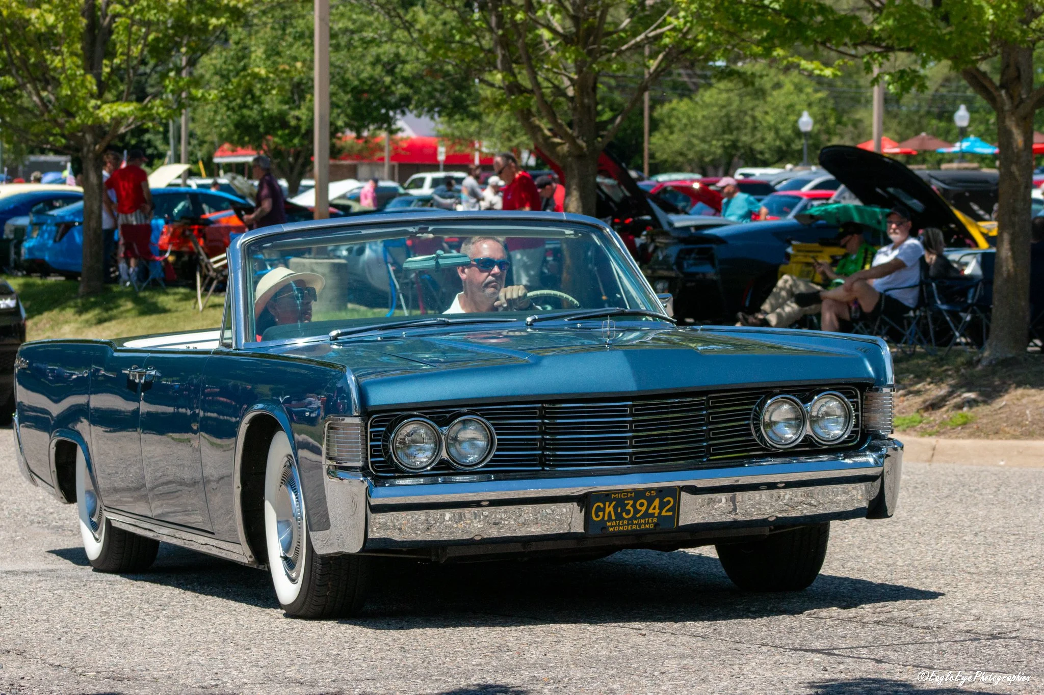 A vintage blue Cadillac driving down a street with people walking and sitting under trees at a car show.