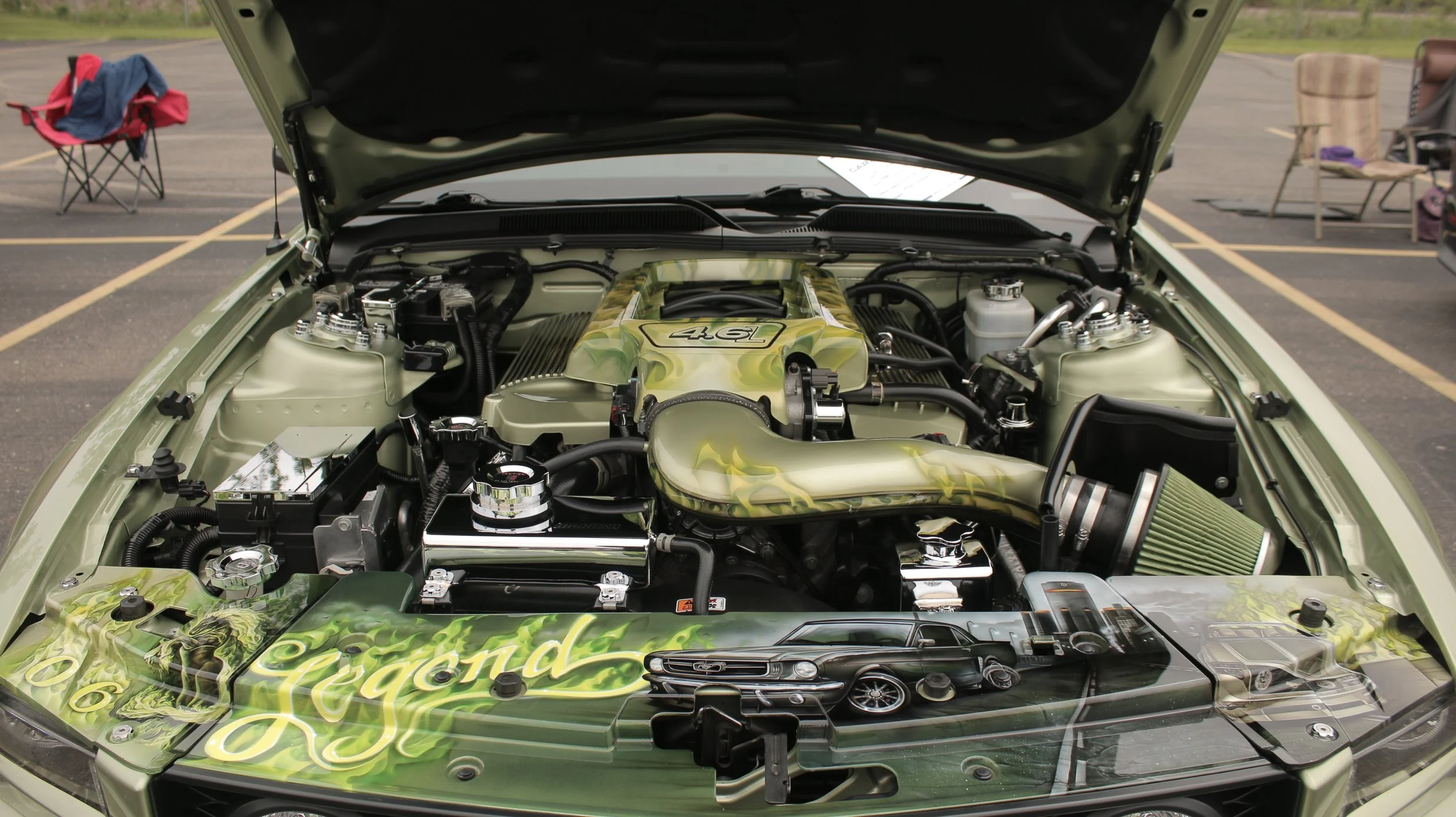 The engine of a car with a custom green paint job and a 4.QLabeled engine cover, parked in an outdoor parking lot with chairs and camping gear in the background.