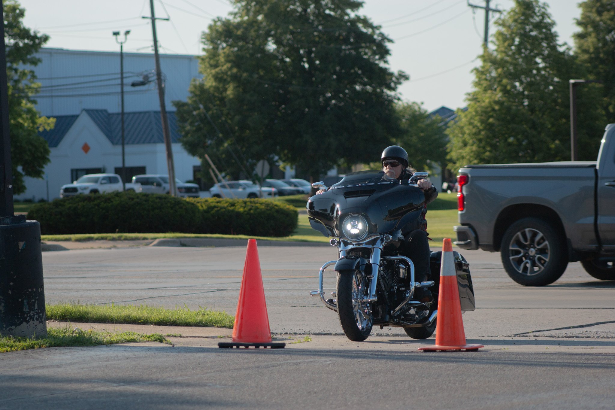 A person wearing a helmet and sunglasses riding a motorcycle on a street with orange traffic cones, with cars and trees in the background.
