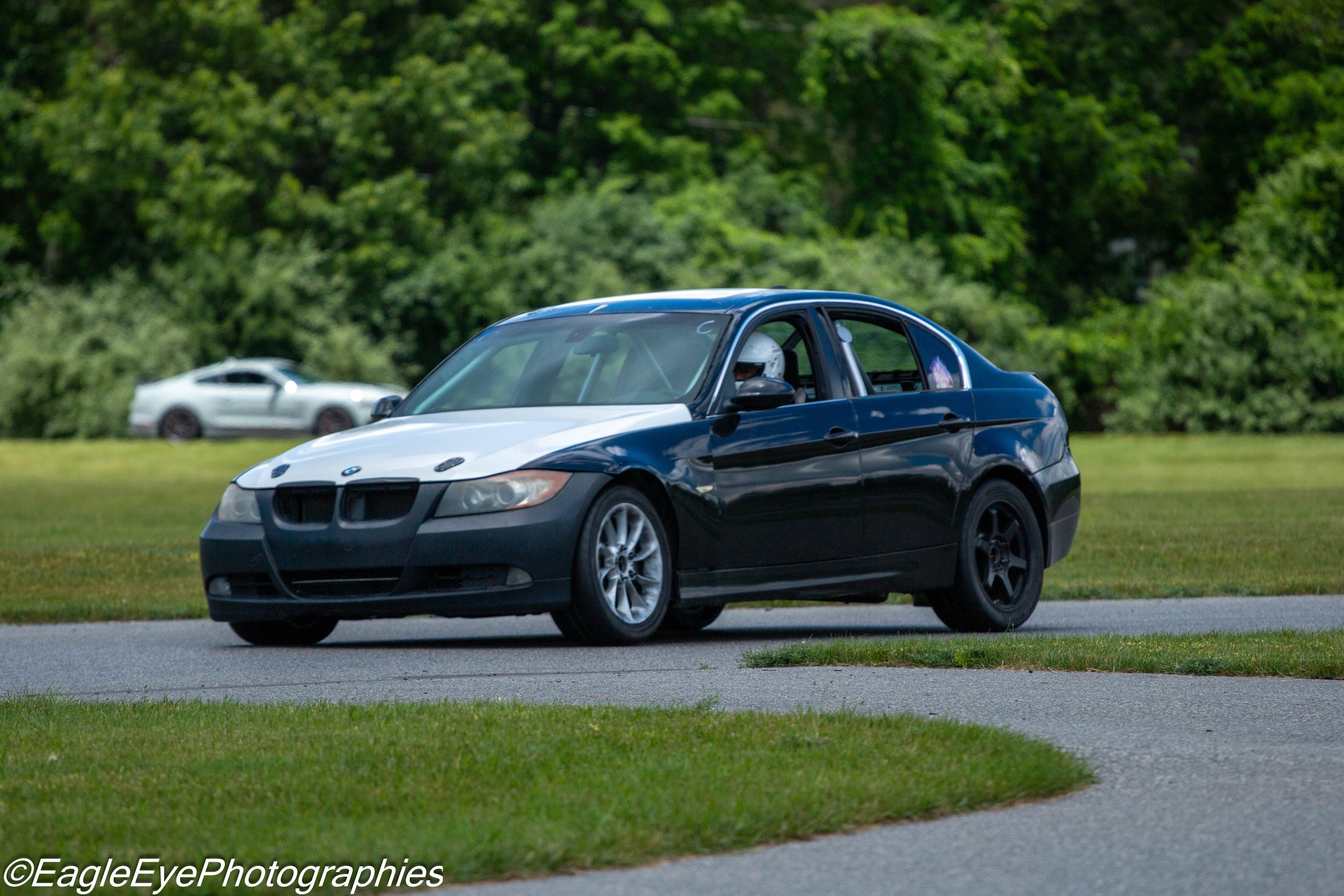 Black BMW sports car with white hood and racing stripes driving on a grassy area, with a silver sports car in the background amidst green trees.