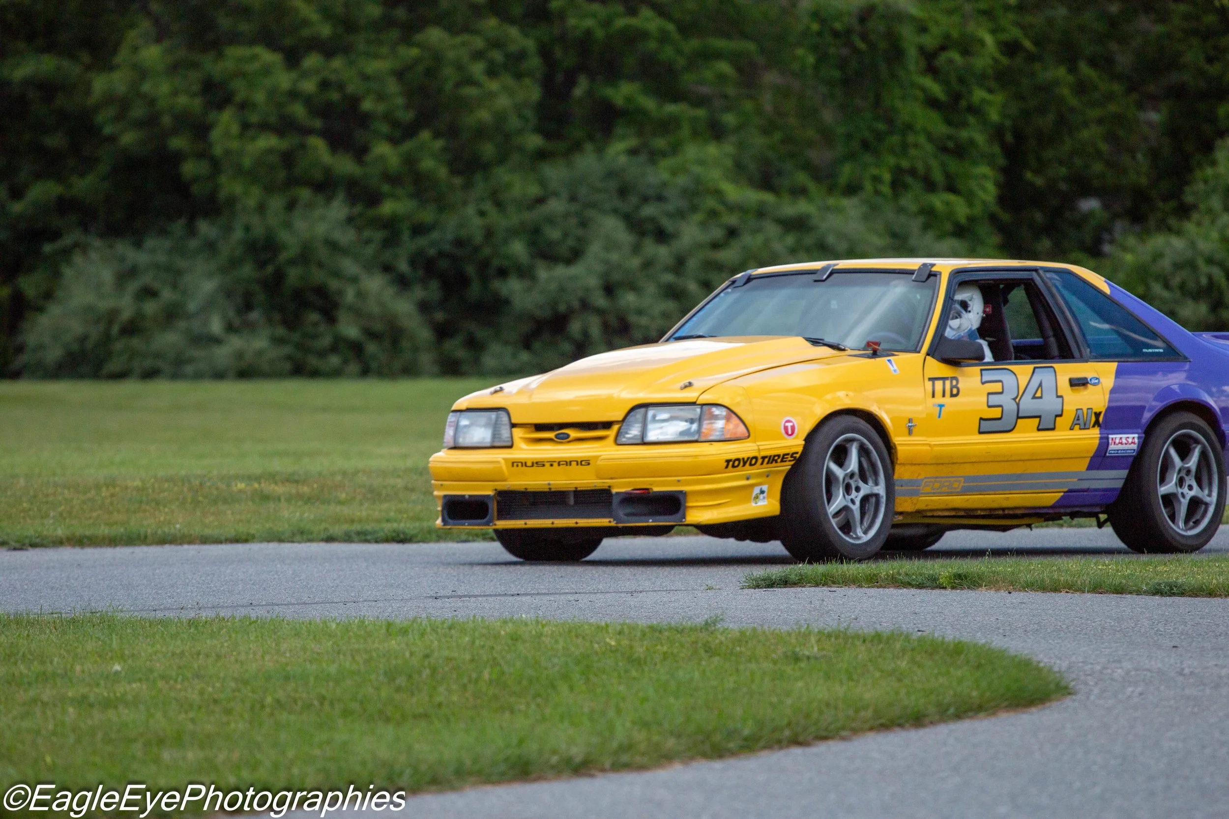 A yellow and blue race car with the number 34 on the side, parked on a grassy area with a background of green trees.