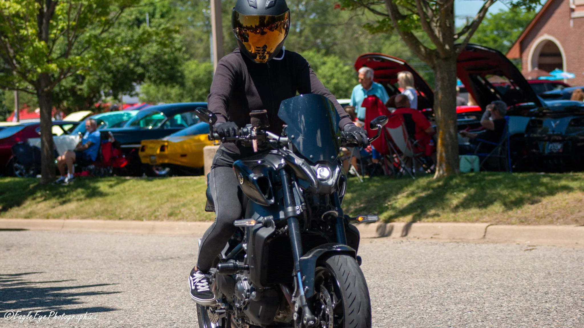 Person wearing a black helmet and black jacket riding a black motorcycle on a sunny day, with a crowd of people and classic cars in the background.