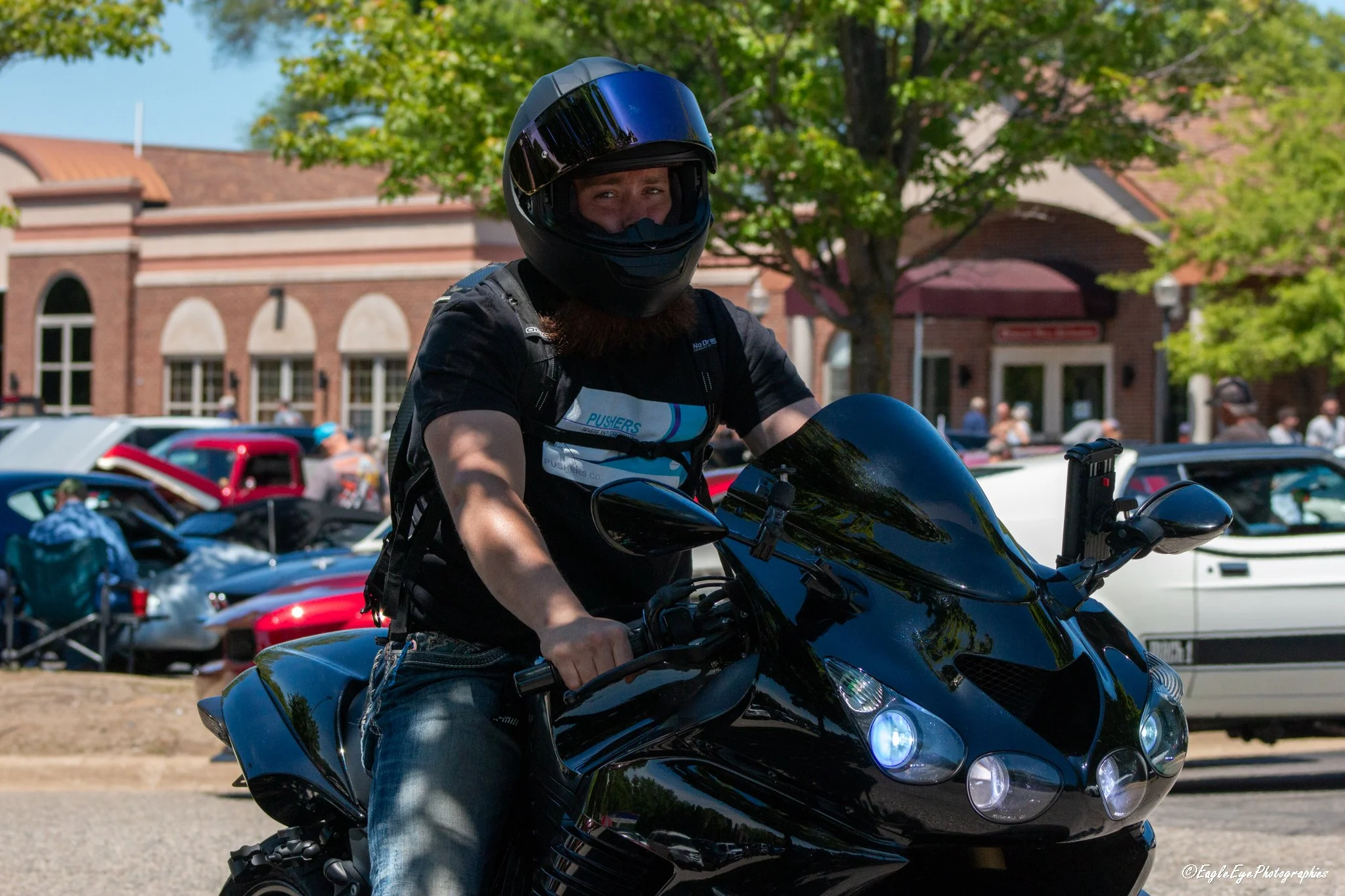 A person wearing a black motorcycle helmet and black shirt riding a black motorcycle in a parking lot with trees and a red brick building in the background.