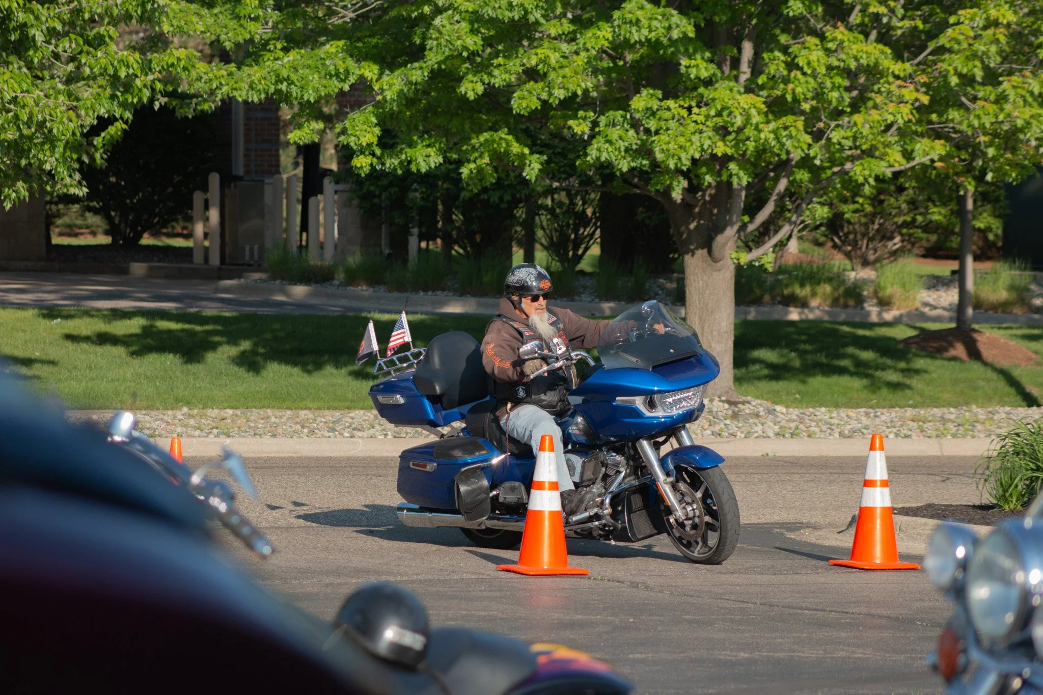 A man wearing a helmet and sunglasses riding a blue motorcycle through a course marked with orange cones, set outdoors with green trees in the background.