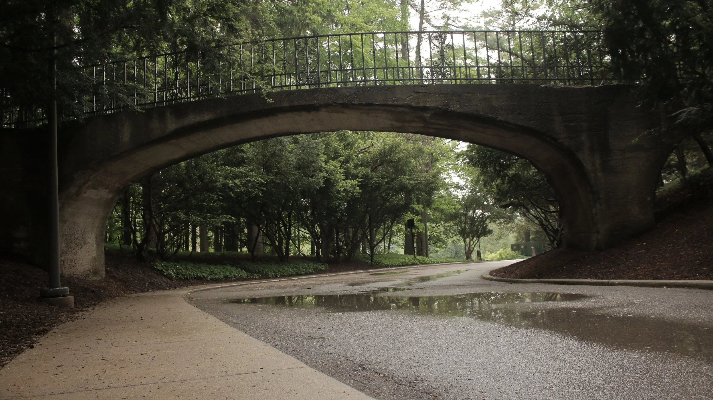 Stone arch bridge over a winding road surrounded by trees and greenery.
