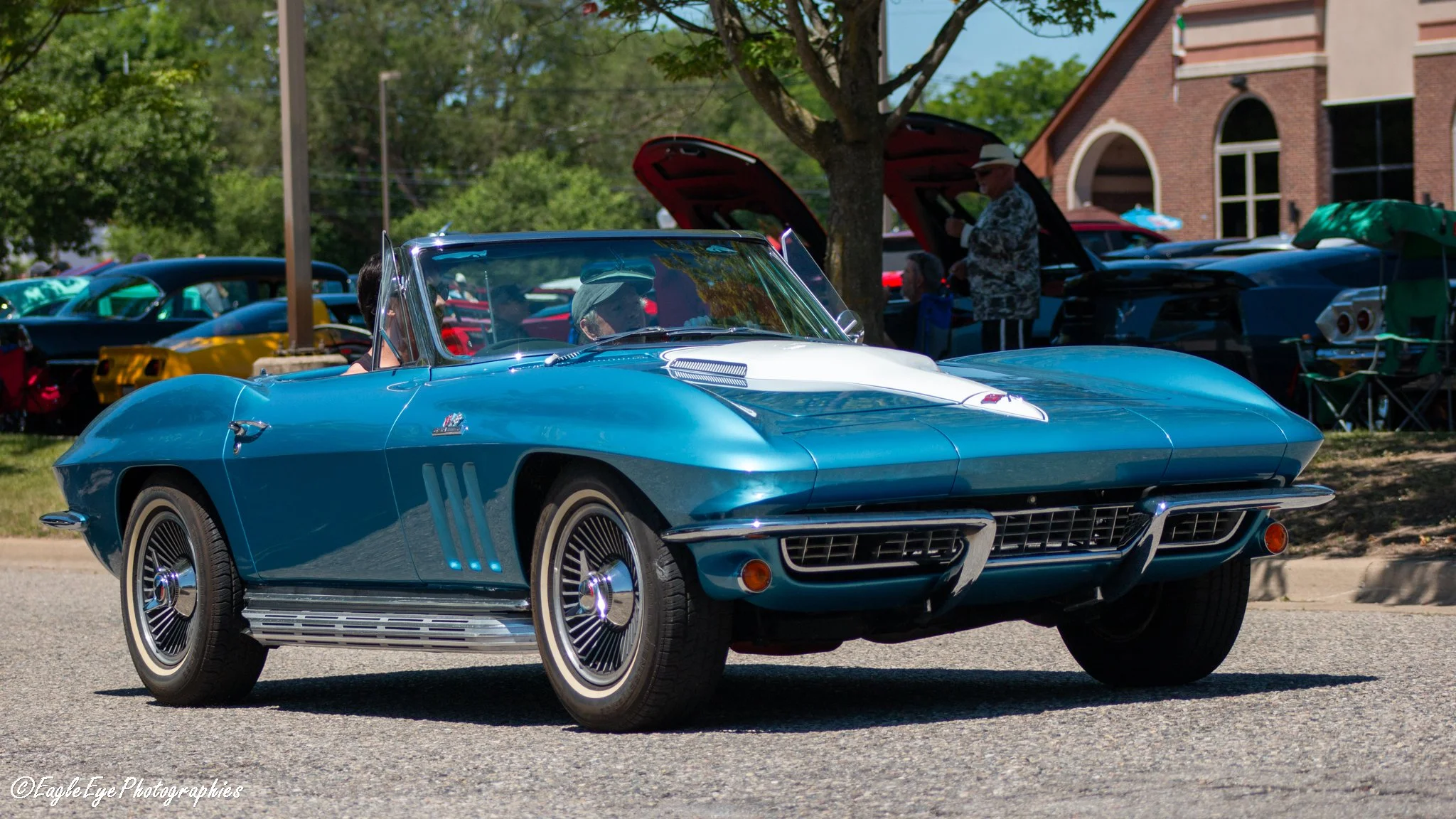 A vintage blue Corvette car parked outdoors at a car show, with other classic cars and people in the background.